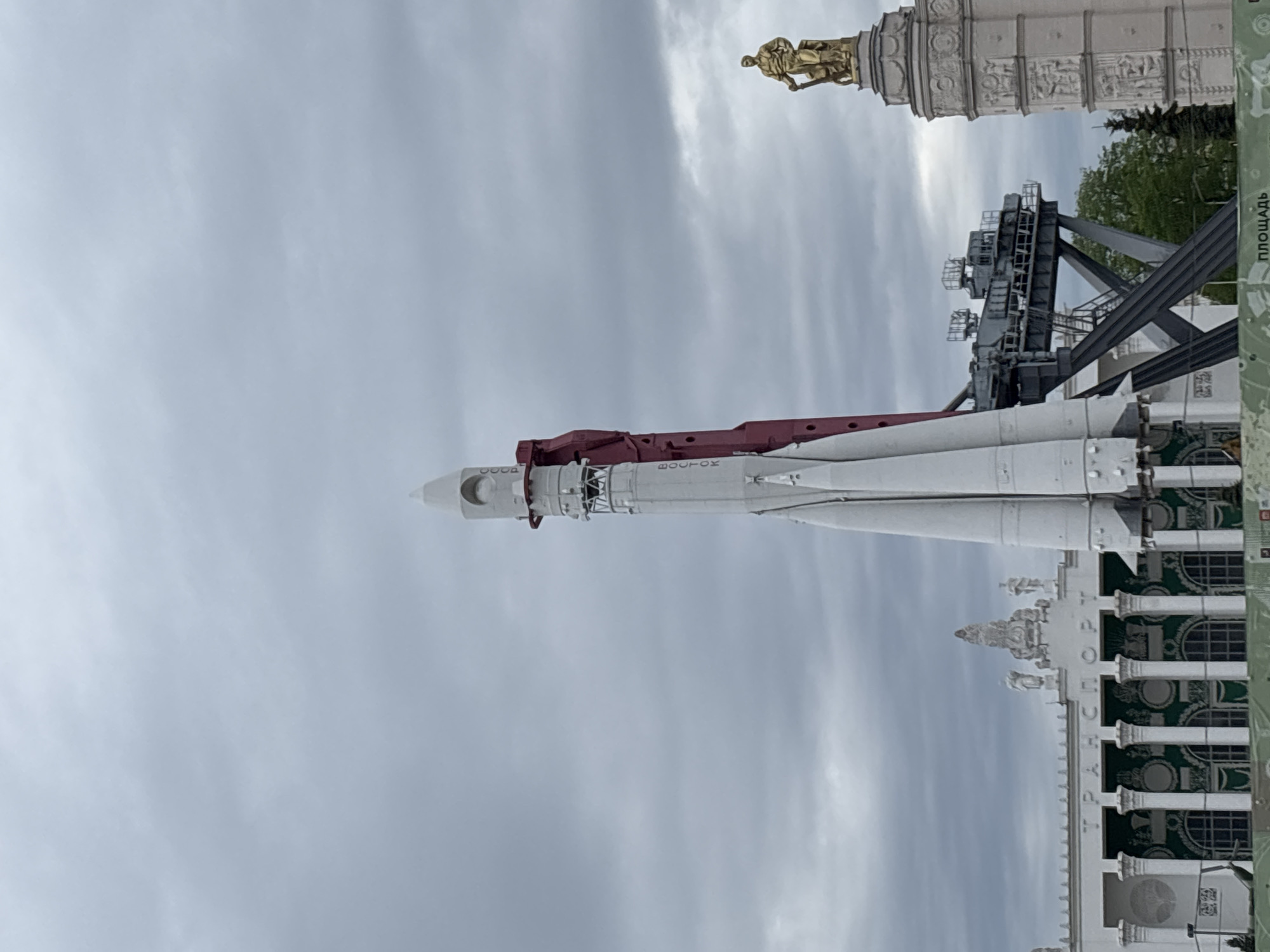 The image shows the Vostok rocket, a Soviet rocket, exhibited at the VDNKh exhibition park in Moscow, Russia. The rocket is white with red and grey sections, oriented horizontally in the frame. Behind the rocket, there's a grey overcast sky. Several buildings are also visible: a section of a white building with green and white designs, a grey service structure, and the upper portion of a columned tower topped with a golden statue.