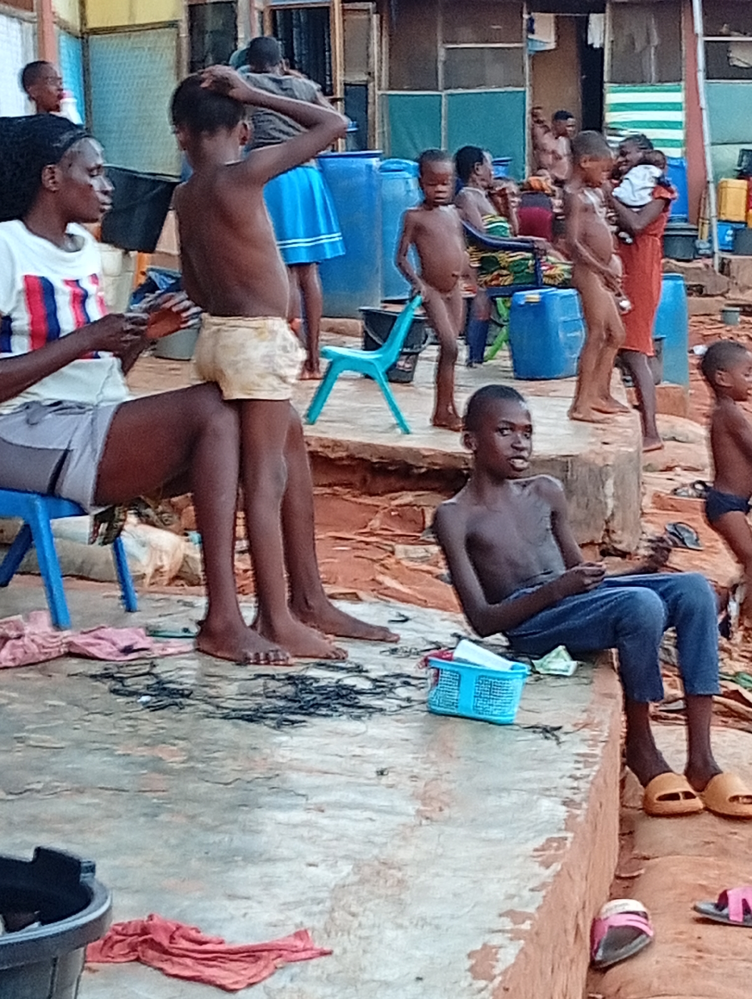 The image depicts a scene in Ogidi, Nigeria. A woman is seated on a blue plastic chair, with a child standing beside her. In the foreground, a shirtless boy sits on a concrete ledge wearing jeans and slippers, holding an object. Several other children are visible in the background, some unclothed or partially clothed. There is a variety of objects in the scene, including blue plastic barrels, a blue plastic basket, clothing, and discarded items scattered on the ground. The environment appears to be an outdoor area with evidence of dwellings in the background.