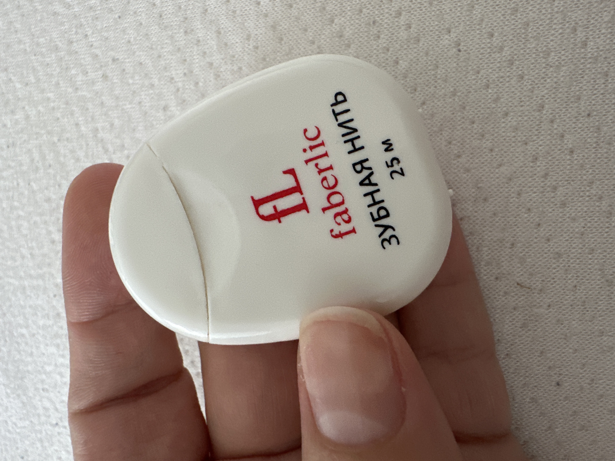 A person's hand holds a small, white, oval-shaped container of Faberlic brand dental floss. The container has red lettering that reads "faberlic" below a red logo that resembles a stylized "FL." Below this, in black lettering, it says "ЗУБНАЯ НИТЬ" (which translates to "dental floss" in Russian) and "25 м" (indicating 25 meters). The background is a textured, light-colored surface, possibly fabric. The image was taken at an unknown location in Krefeld, Germany.