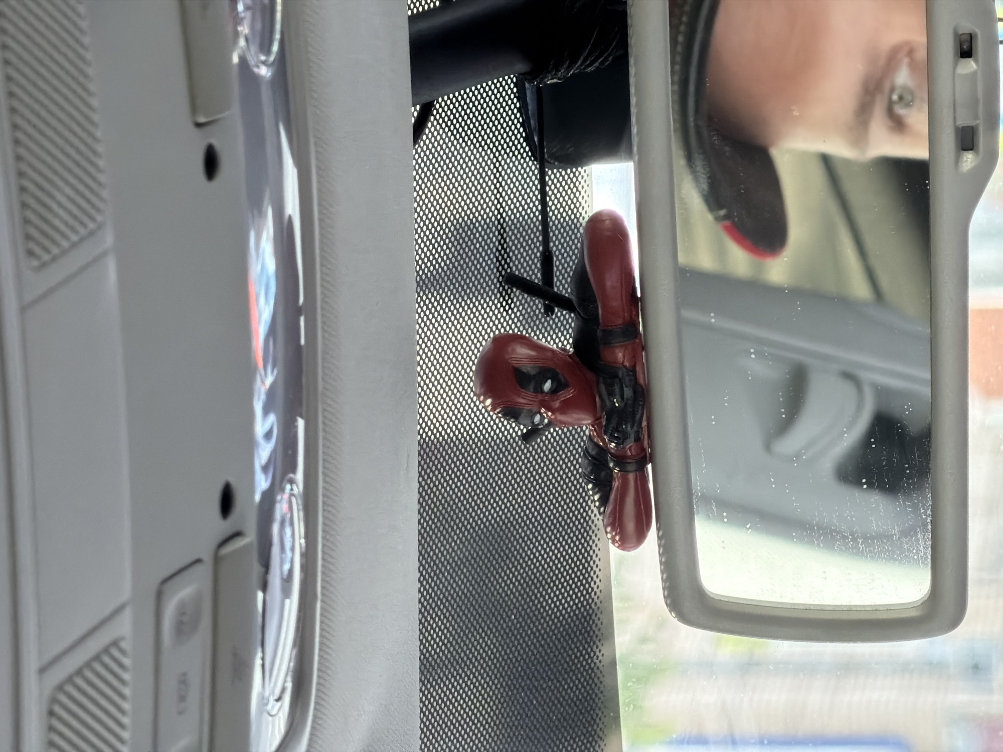 The image shows the interior of a vehicle, likely a car. A Deadpool figurine is hanging from the rearview mirror. The mirror reflects a partial image of a person with a piercing. The car's interior details include a textured headliner and controls near the roof. The car's location appears to be in the city of Chaikovsky, Russia, based on information provided.