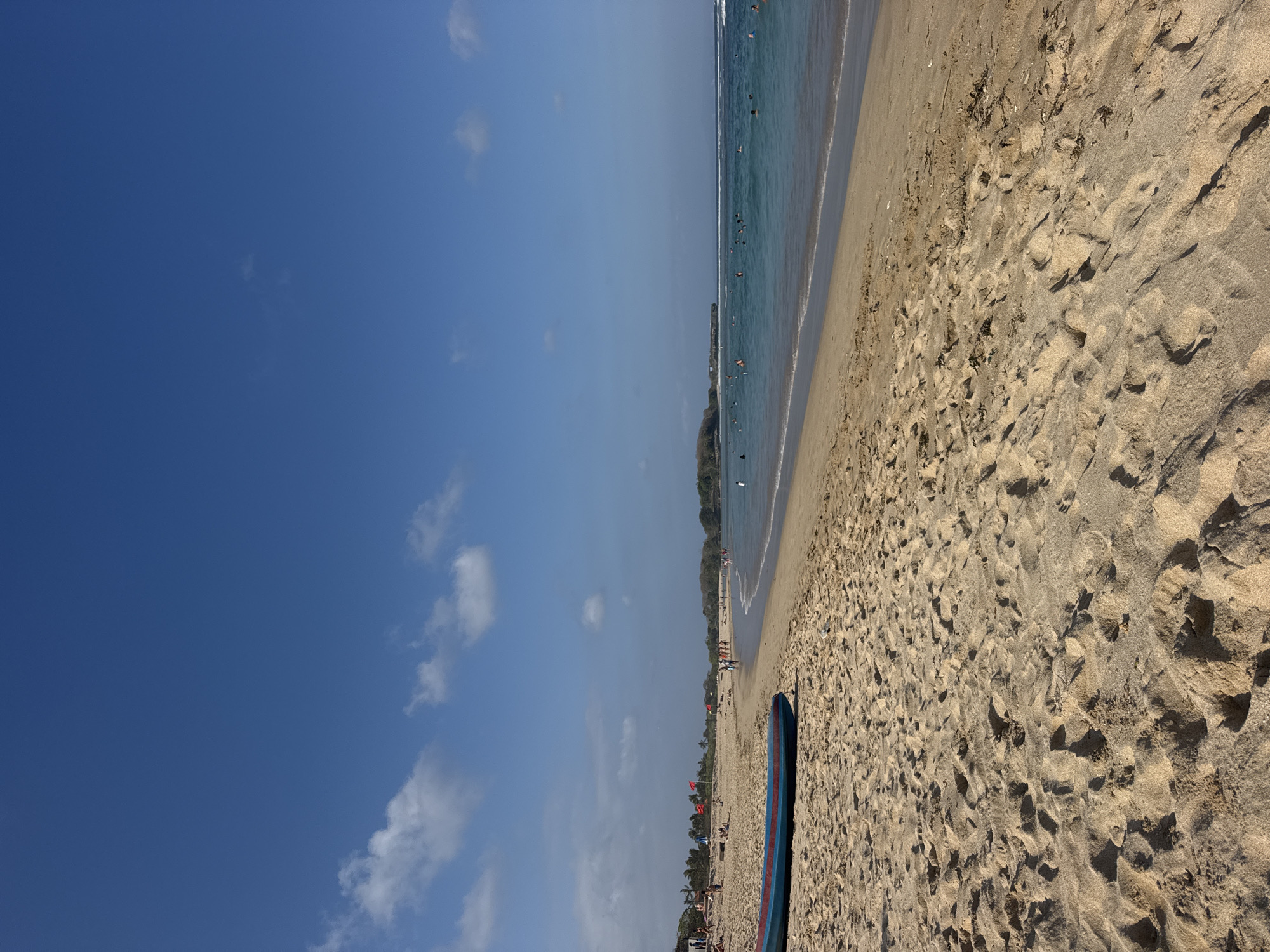 The image depicts a sunny day at Kuta Selatan beach in Indonesia. The foreground is dominated by sand with footprints. A long, narrow boat, blue and red, rests on the sand. In the midground, a sandy beach stretches out, populated by numerous people, some walking and others relaxing. Red flags are visible on the beach. Further out, the ocean is blue with moderate waves, and many people are swimming or surfing. The horizon shows a line of trees and land, under a clear blue sky with scattered white clouds.