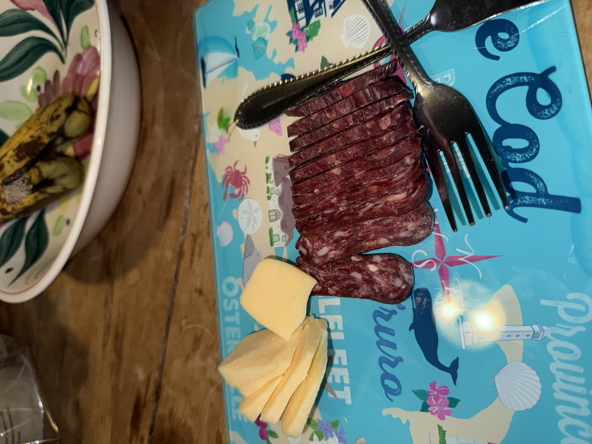 The image shows a charcuterie board with sliced sausage and cheese, arranged with a knife and fork on a Provincetown-themed cutting board. A bowl with overripe bananas sits to the left. The scene is set indoors on a wooden surface.