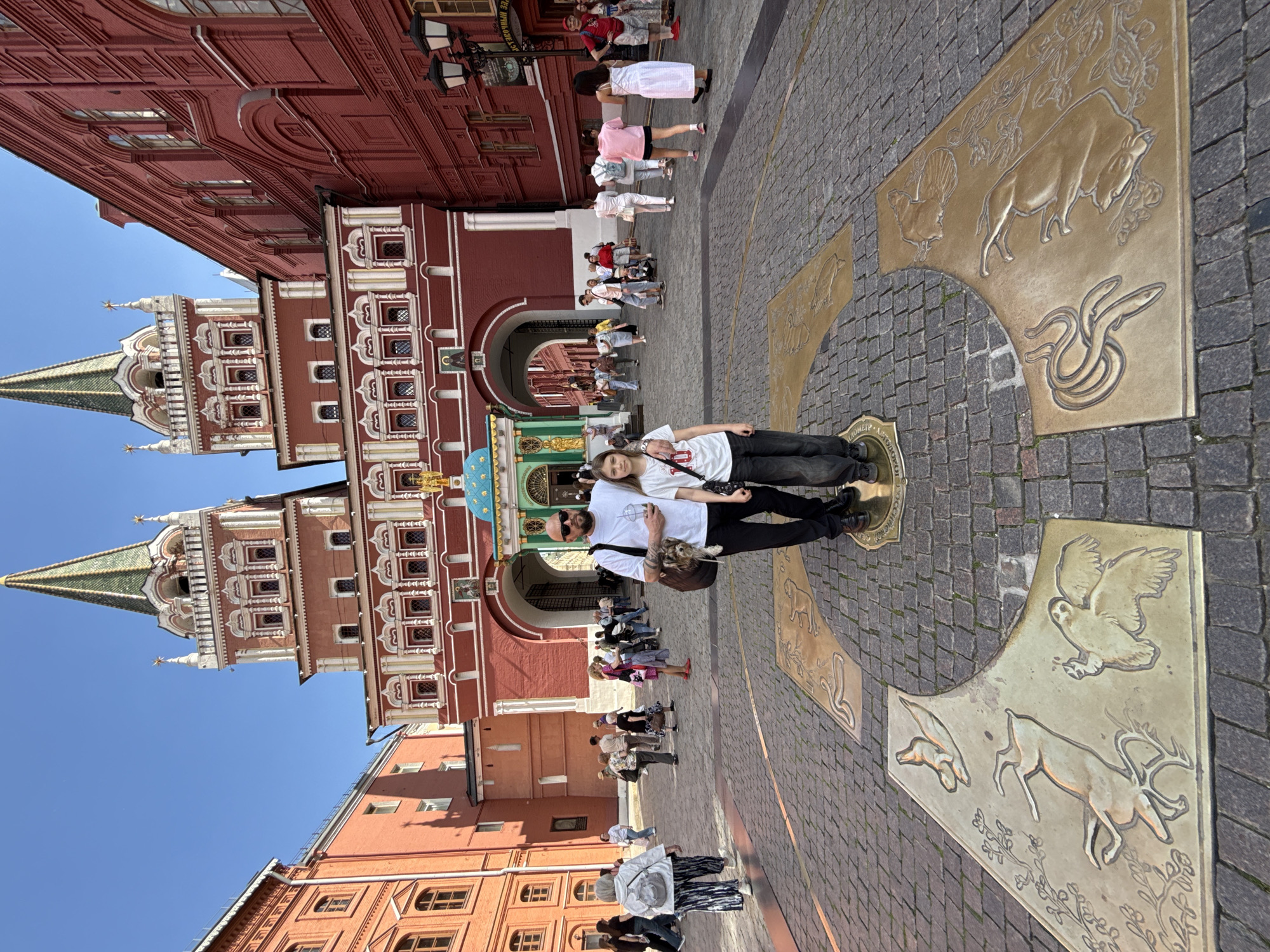 The image displays a vibrant outdoor scene in a historic city square, identified as Moscow, Russia, under a clear blue sky. Bright sunlight casts distinct shadows, suggesting it is likely late morning or early afternoon.
In the foreground, an adult male and a younger female stand centrally on a large, ornate brass medallion embedded in the cobblestone pavement. The medallion is richly decorated with relief carvings of various animals, including a bull, rooster, snake, deer, and an owl, interspersed with floral patterns. Text visible on the central part of the medallion reads "НУЛЕВОЙ КИЛОМЕТР," which translates to "ZERO KILOMETER," a known landmark in Moscow.
Behind the two individuals, a grand, red-brick building dominates the left side of the frame. It features elaborate white decorative trim around its numerous windows and cornices, and is crowned by two prominent towers with pointed green spires. An arched passageway cuts through the base of this building. Beyond this and further into the square, many other people are visible, walking leisurely or standing in groups, indicating a public and likely tourist-frequented area. The overall activity suggests sightseeing and casual strolling in a lively urban setting.