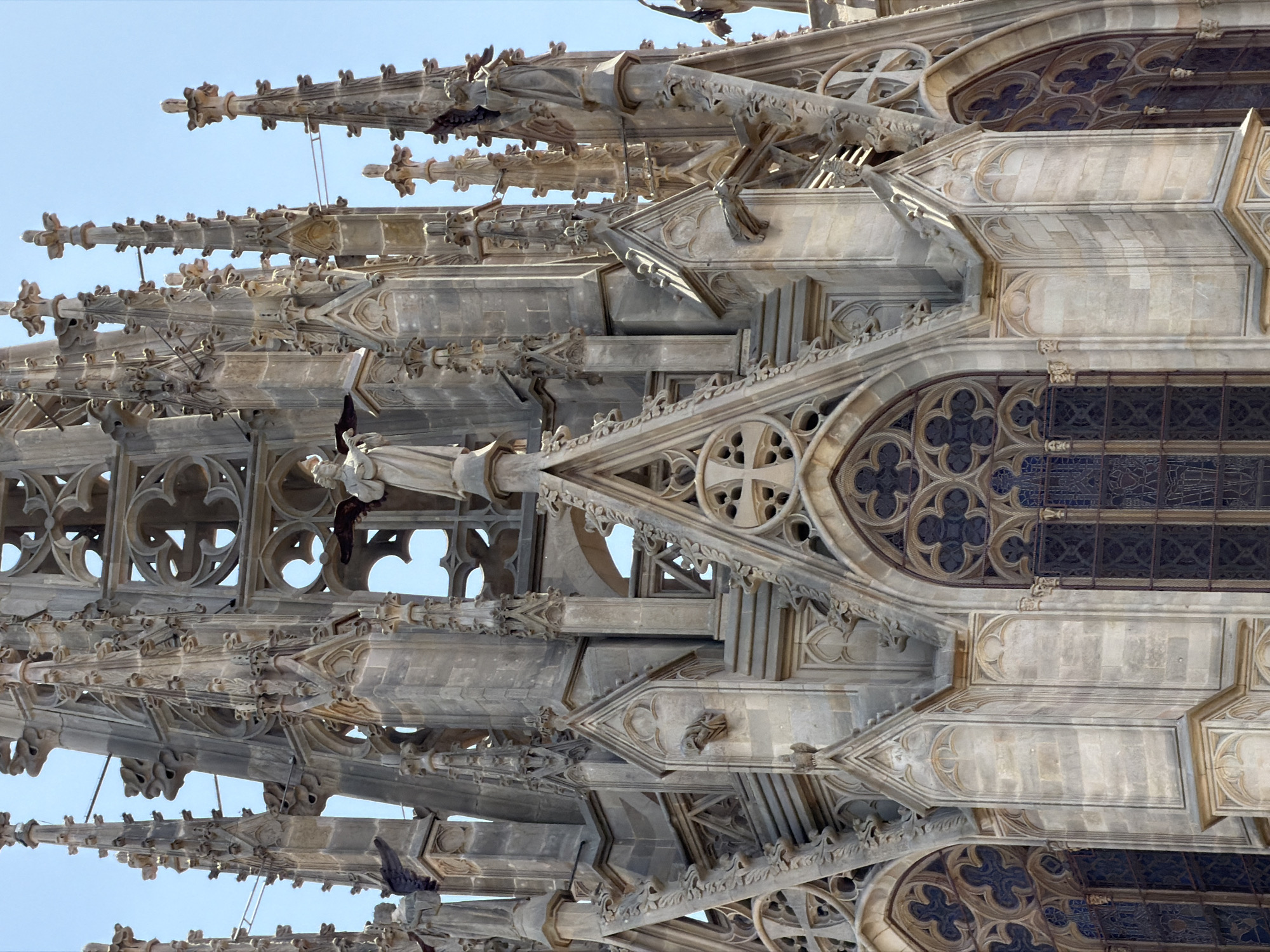 The image shows a close-up, low-angle view of the spires and upper facade of the Barcelona Cathedral, located in the Gothic Quarter of Barcelona, Spain. The architecture is characterized by intricate Gothic detailing, including spires, pinnacles, flying buttresses, and ornamental tracery. Several statues of figures, possibly saints or angels, are visible in niches and on ledges. The stonework appears weathered, with a greyish-brown hue, and the sky is a clear, pale blue. The windows are tall and narrow, with dark panes and decorative tracery. Gargoyles are visible protruding from the stonework. The overall impression is one of grand, elaborate religious architecture.