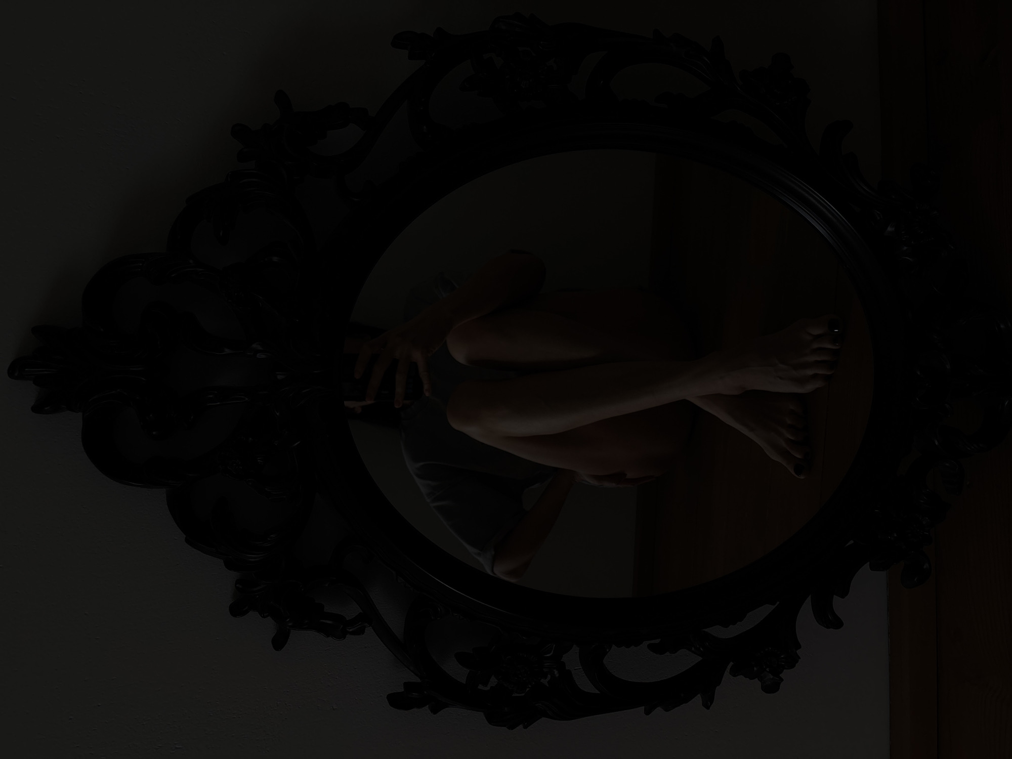 A dim, low-light photograph captures a dark, ornate-framed round mirror hanging on a wall. The mirror reflects a person seated with their arms crossed and head down, obscuring their face. The frame is black and features elaborate floral and scroll designs. The wall behind the mirror is plain and dimly lit. The reflection suggests a dark or enclosed space. There is no information to suggest this image was taken in Chaikovsky, Russia.
