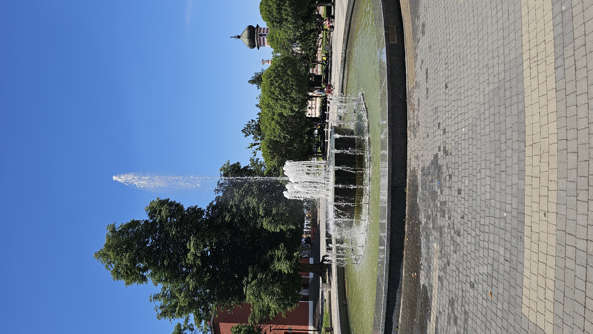 The image captures a park scene in Unknown, Unknown, featuring a tiered fountain as the central element. The fountain is positioned in a circular pool filled with greenish water. A single, high jet of water shoots up from the center, contrasting with the clear blue sky. Trees are adjacent to the fountain, obscuring some of the building in the background. A building with a green dome is located on the center. The ground is paved with interlocking stones in alternating shades of gray and tan.