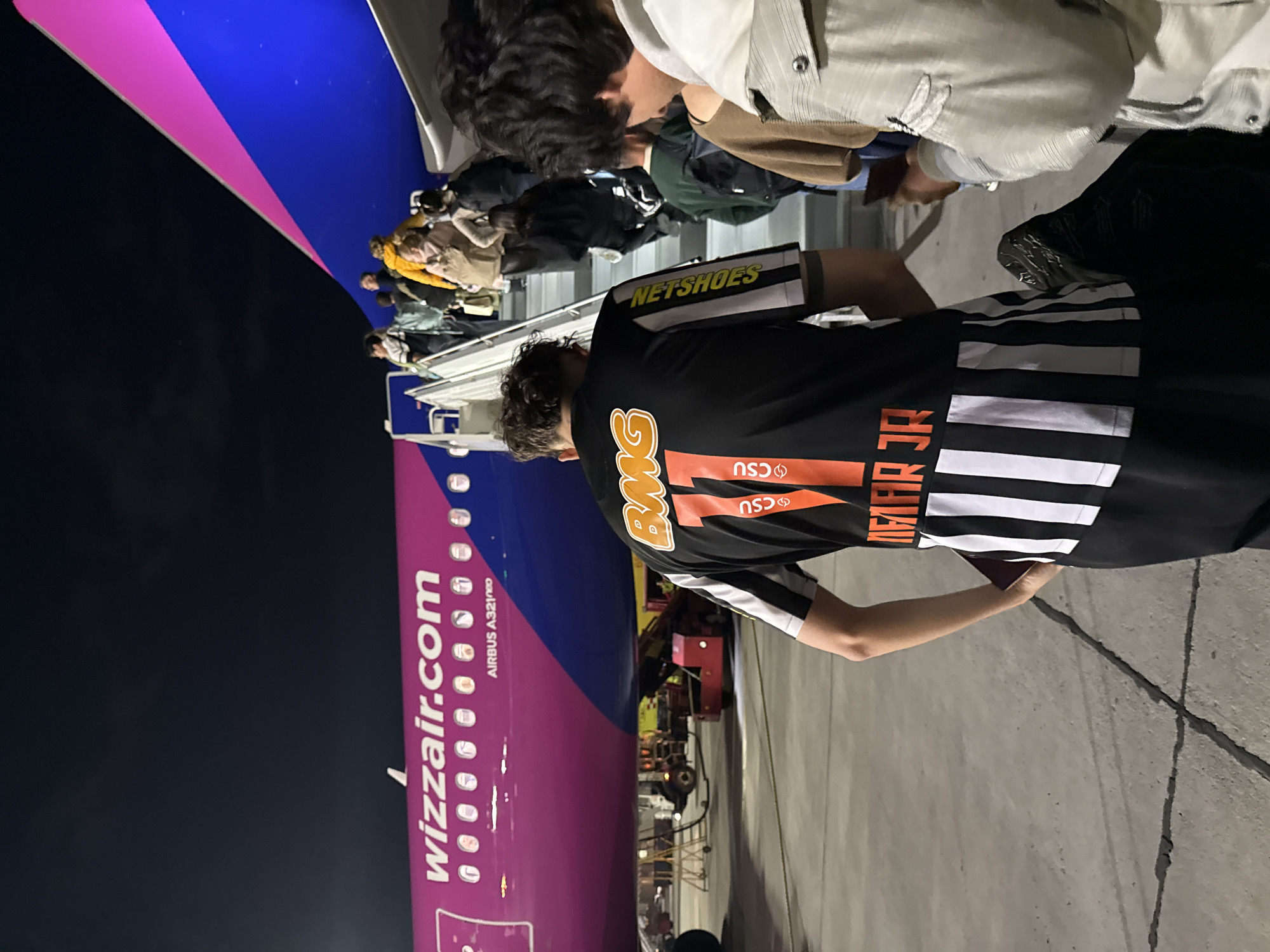 The image depicts a nighttime scene at an airport, likely Zvartnots International Airport in Yerevan, Armenia, as passengers board a Wizz Air aircraft.
In the foreground, the back of a person is visible, wearing a black shirt with prominent orange and white details. Large orange letters spelling "BMG" (or "PMG") and the number "7" are displayed on the back. Below this, the letters "CSU" appear in orange, potentially repeated. The right sleeve of the shirt features yellow text that reads "NETSHOES", and the lower back of the shirt has several horizontal white stripes. This individual is holding a small, dark object in their left hand, possibly a passport or phone.
Further ahead, a line of passengers is seen ascending mobile boarding stairs or a jet bridge to enter the aircraft. They are casually dressed, with one person in a yellow jacket visible among them.
The left side of the frame is dominated by the fuselage of a Wizz Air Airbus A321neo airplane. Its distinctive purple and blue livery is clearly visible. White lettering on the purple section spells out "wizzair.com", and below it, "AIRBUS A321neo" is printed.
The scene is illuminated by artificial lighting from the airport, indicating it is nighttime. The ground appears to be a dark concrete or asphalt tarmac with visible cracks. The sky is dark, and no specific weather conditions are discernible beyond a clear night. The overall impression is that of a standard commercial flight boarding process.