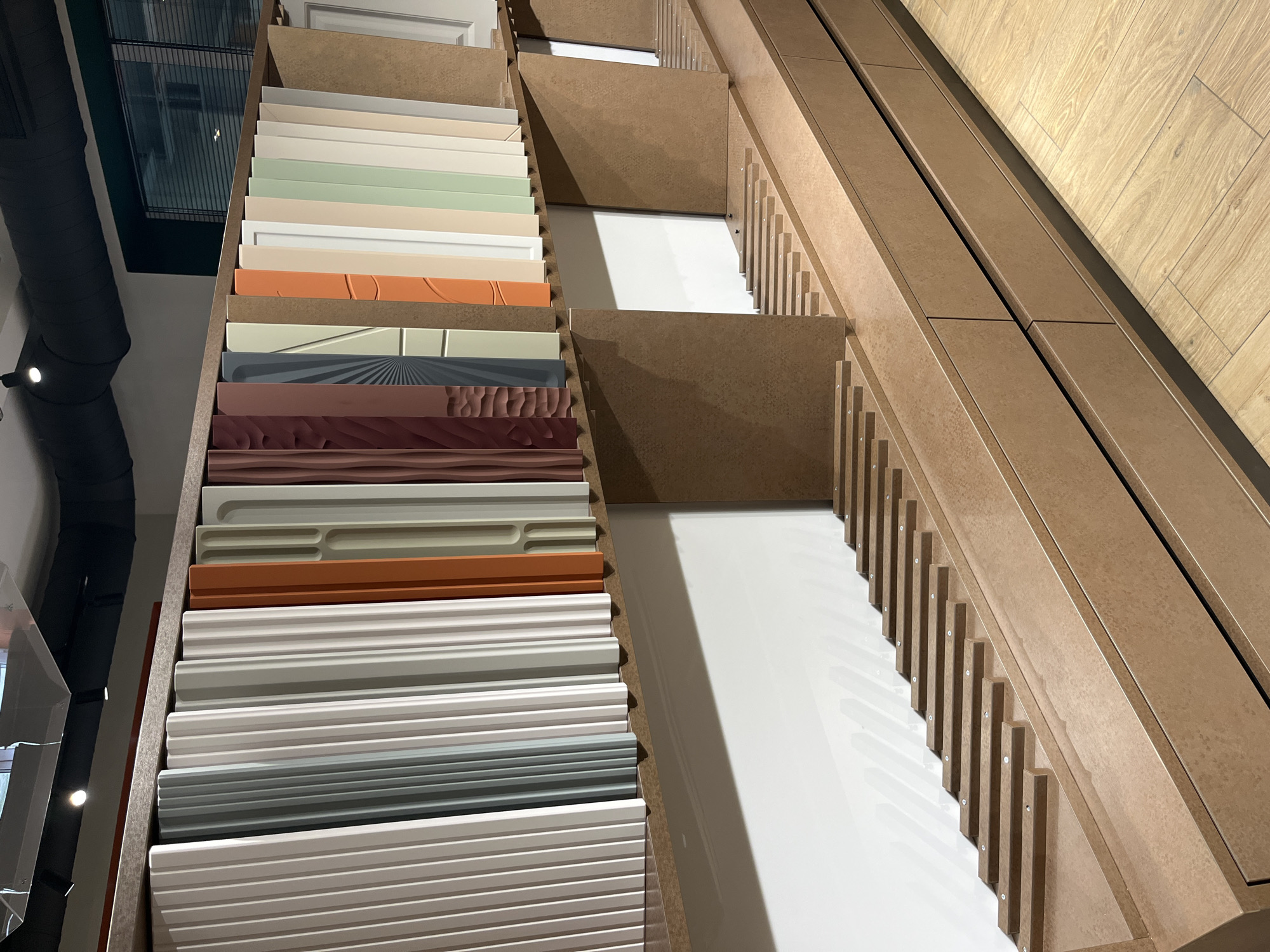 The image presents a display of various surface samples, likely for interior design or construction purposes. The samples are arranged in tiered shelves, with the shelves constructed from a light brown, possibly MDF, material. On the left side, the samples are visible in full, showcasing different colors, textures, and patterns. There are creams, greens, oranges, and several shades of grey and white, including some with embossed designs. The right side shows larger, white samples with only the edges of the shelves visible, and there are rows of vertical supports along the sides. The background of the right side has a light-colored, wood-grained surface. The lighting is bright and even, casting soft shadows. The overall setting seems to be an indoor showroom or design studio. The location is Unknown, Unknown. There are no people visible.