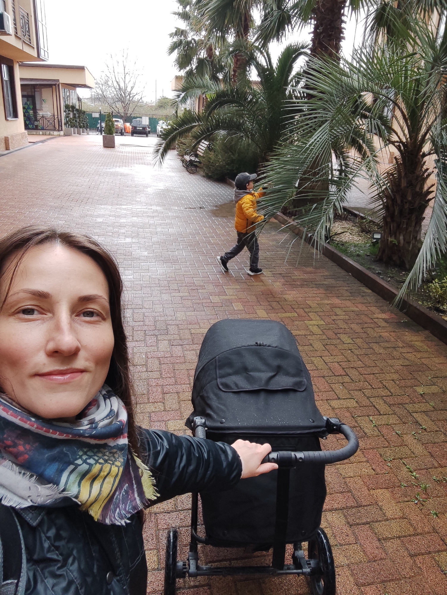 The image shows a woman taking a selfie while pushing a black stroller along a brick-paved walkway. A child in a yellow jacket and grey cap walks ahead, near some palm trees. The walkway is lined with buildings on one side and palm trees on the other. The scene is set in Sochi, Russia. The ground appears slightly wet, suggesting it may have recently rained.