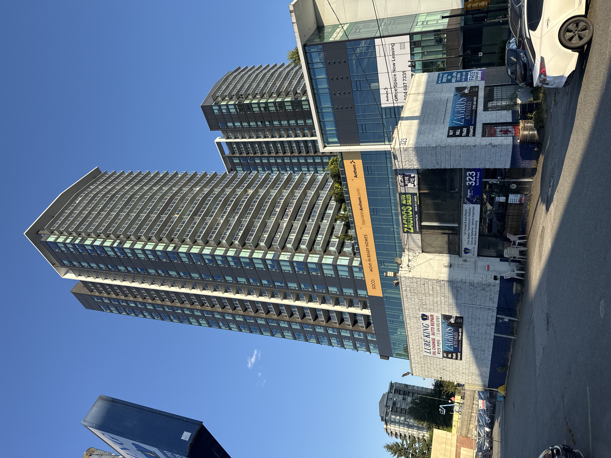 The image depicts a street-level view of a commercial building facade and a tall residential tower in Burnaby, Canada. The commercial building, located at 323 North Road, features a white brick facade with various signage for businesses, including "LUBE KING OIL CHANGE AUTO REPAIR," "ZAGROS AUTO SALES," and "DENTAL SPA." Above this building, a modern glass-fronted structure is visible, advertising "Office Space Now Leasing" by Anthem. Towering behind these structures is a large, dark-colored residential high-rise with numerous balconies. The sky is clear and blue, indicating a daytime scene. Several parked cars are visible along the street and in a lot adjacent to the commercial building. The overall scene suggests an urban environment with a mix of residential and commercial properties.