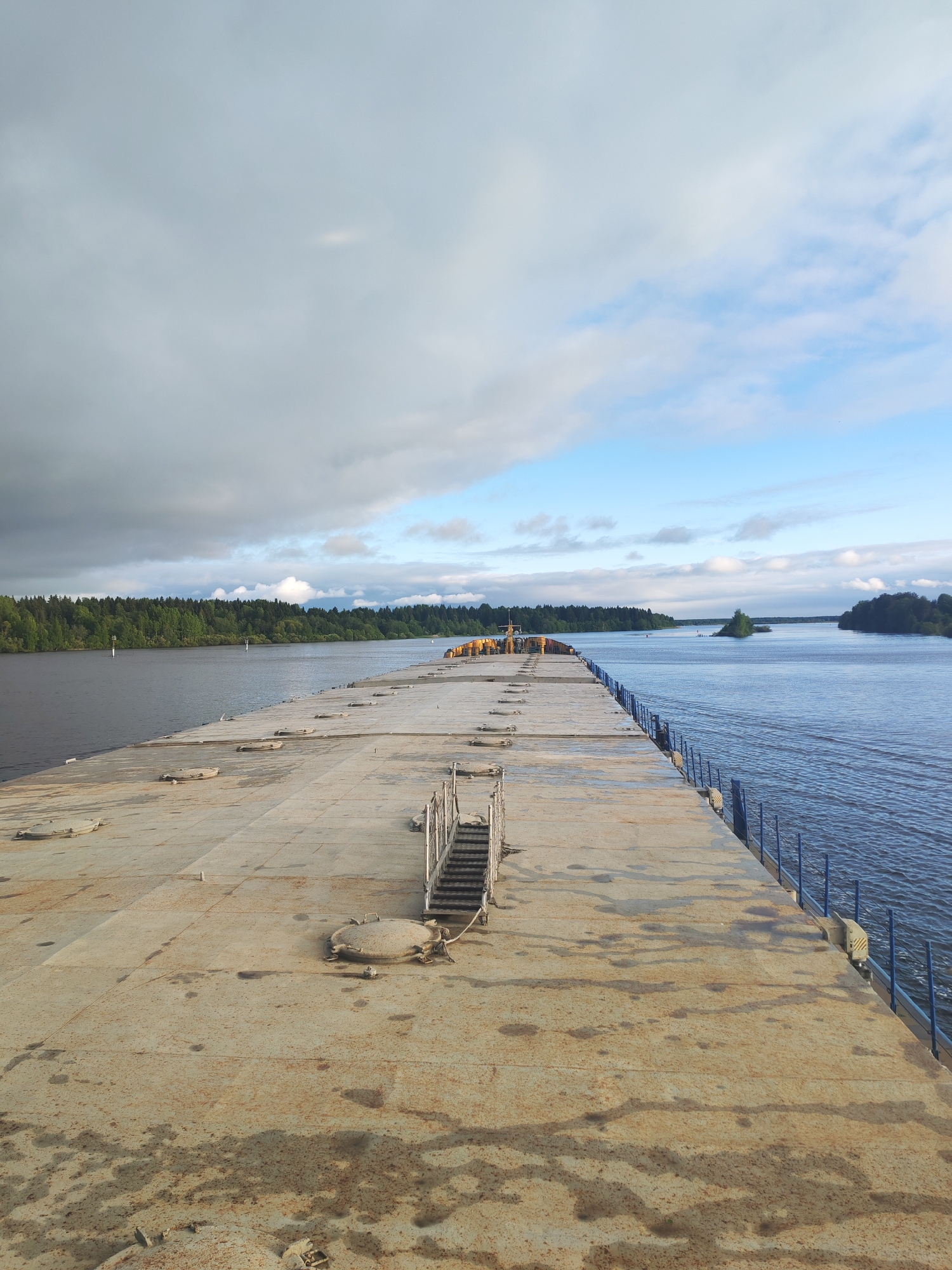 The image depicts a long, flat, weathered metal surface, possibly a barge or dock, extending into a body of water. The metal surface shows signs of rust and staining. A set of metal stairs with railings is positioned towards the center of the frame. A railing with blue vertical supports runs along the right edge of the metal structure.
The water is calm with some visible ripples. In the distance, the shoreline is lined with green trees. There is an island to the right. The sky is overcast with a mix of gray and blue clouds.
The image does not contain enough information to determine the city and state in which it was taken.