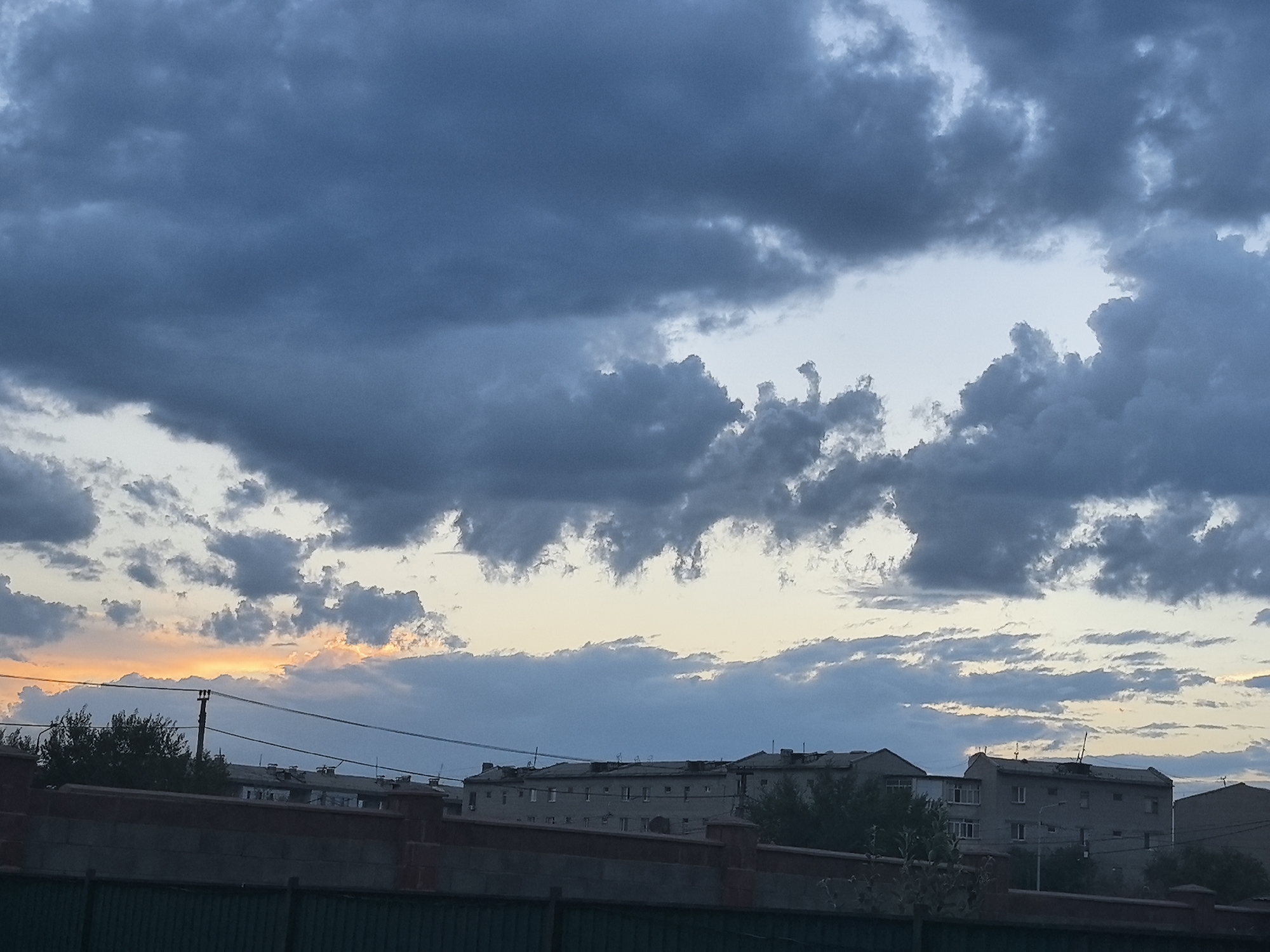 This is a landscape photograph taken in Taldykorgan, Kazakhstan. The image captures a view of buildings and a fence in the foreground, with a dramatic sky filled with clouds dominating the upper portion of the frame.
The foreground consists of a low wall made of brick and a dark green fence, suggesting a boundary or property line. Behind this, several multi-story buildings are visible. These buildings appear to be residential or apartment complexes, with windows and architectural details characteristic of urban environments. Trees and some vegetation are scattered between and in front of the buildings, adding a touch of nature to the scene. A utility pole with wires crosses the frame horizontally, indicating the presence of infrastructure.
The sky is the most prominent feature of the image. It is filled with a variety of clouds, ranging from dark, heavy formations to lighter, more wispy ones. Towards the left side of the frame, a band of warm, orange and yellow light streaks across the horizon, indicating either a sunrise or sunset. The contrast between the dark clouds and the bright, colorful horizon creates a sense of depth and drama. The overall weather appears to be changeable, with the presence of both dark, potentially storm-laden clouds and pockets of clear or brightly lit sky.
The time of day is either early morning or late evening, given the coloration of the sky. The location cues from the architecture and the general landscape suggest an urban or semi-urban setting in Kazakhstan, specifically Taldykorgan. There is no visible text in the image. The mood of the image is somewhat somber due to the dark clouds, but the colorful horizon adds a hopeful or peaceful element.