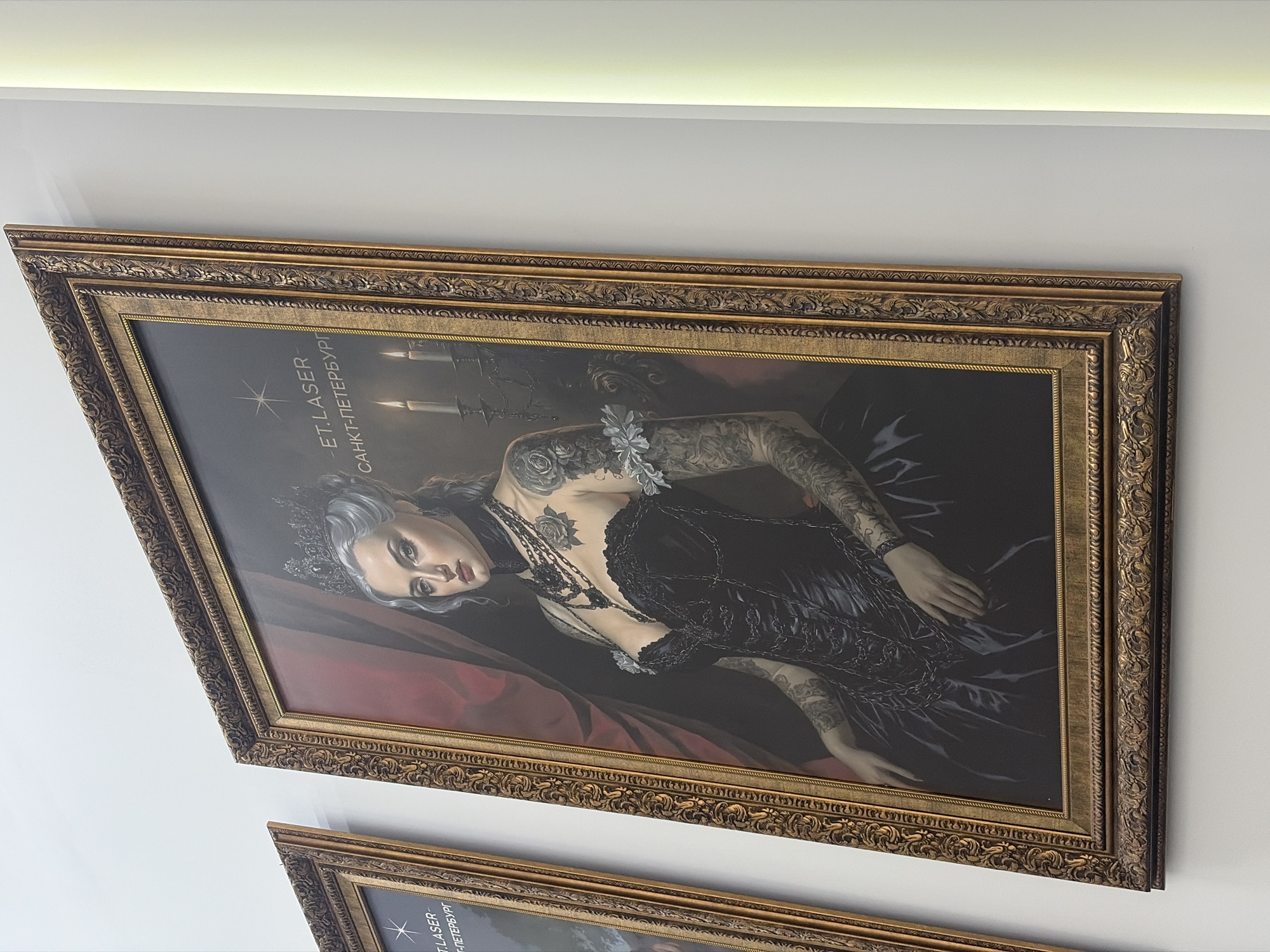 The image displays a framed painting hanging on a light-colored, possibly white, wall, with a partial view of a second similar painting below it. The photograph is taken from an angled perspective, showing the top painting prominently.
The main painting features a portrait of a woman with pale skin, greyish-white hair, and a small dark crown. She has a serious, intense expression with dark lips and eyes. Her arms and upper chest are extensively covered in dark tattoos, depicting floral and possibly other patterns. She is adorned with multiple dark necklaces and appears to be wearing a dark, intricately detailed dress or gown, possibly black, which includes decorative chains. A light-colored, perhaps white, floral embellishment rests near her shoulder. The background of the portrait includes dark red drapery on the left and dimly lit candles in a dark setting on the upper right, suggesting a dramatic or gothic aesthetic.
On the painting, there is vertical text in the upper left corner. It reads "ET.LASER" followed by a star-like symbol, and then "САНКТ-ПЕТЕРБУРГ" (Sankt-Peterburg), which translates to Saint Petersburg.
The frame of the painting is ornate and golden, with intricate carvings and decorative elements, giving it a classic and luxurious appearance. The wall above the painting is illuminated by a hidden light source, likely an LED strip, running along the ceiling line. The overall scene suggests a contemporary setting showcasing art with a dramatic, stylized theme, possibly in a business or gallery in Saint Petersburg, Russia.