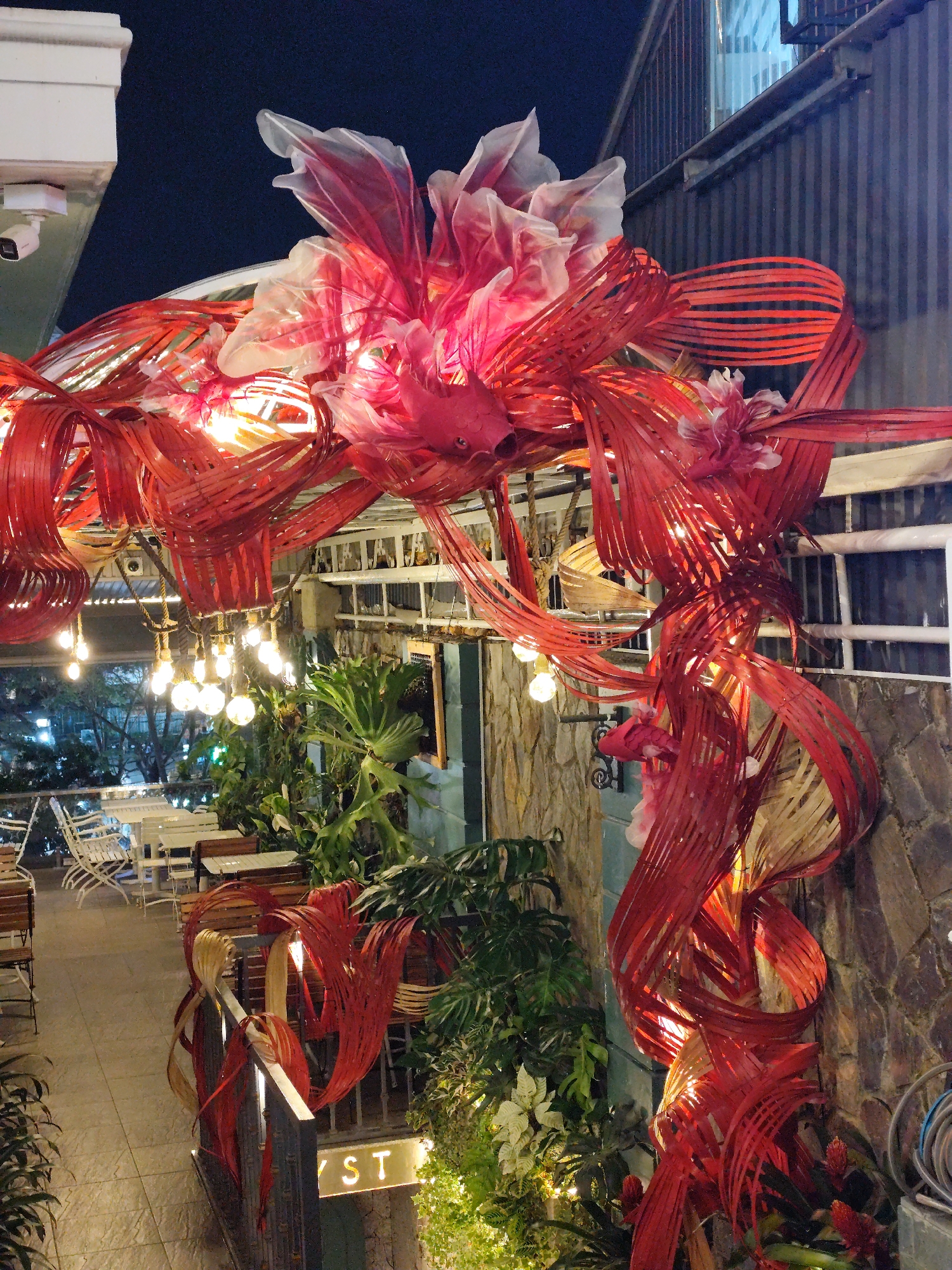 This photograph captures an outdoor scene in Da Lat, Vietnam, where the entrance to a restaurant or venue is adorned with elaborate red and white decorations, possibly for a special occasion or holiday. The decorations consist of strips of red material shaped into swirls and floral arrangements. Illuminated string lights are draped around the decorations and plants, adding to the warm ambiance. The decor is mounted on a stone wall. In the background, there are tables and chairs on a patio, suggesting an outdoor seating area for the restaurant. The surroundings include greenery and architectural elements of the building, such as windows, railings, and a security camera. At the base of the decoration is a sign that reads "ST". The lighting and time of day suggest it is evening.
