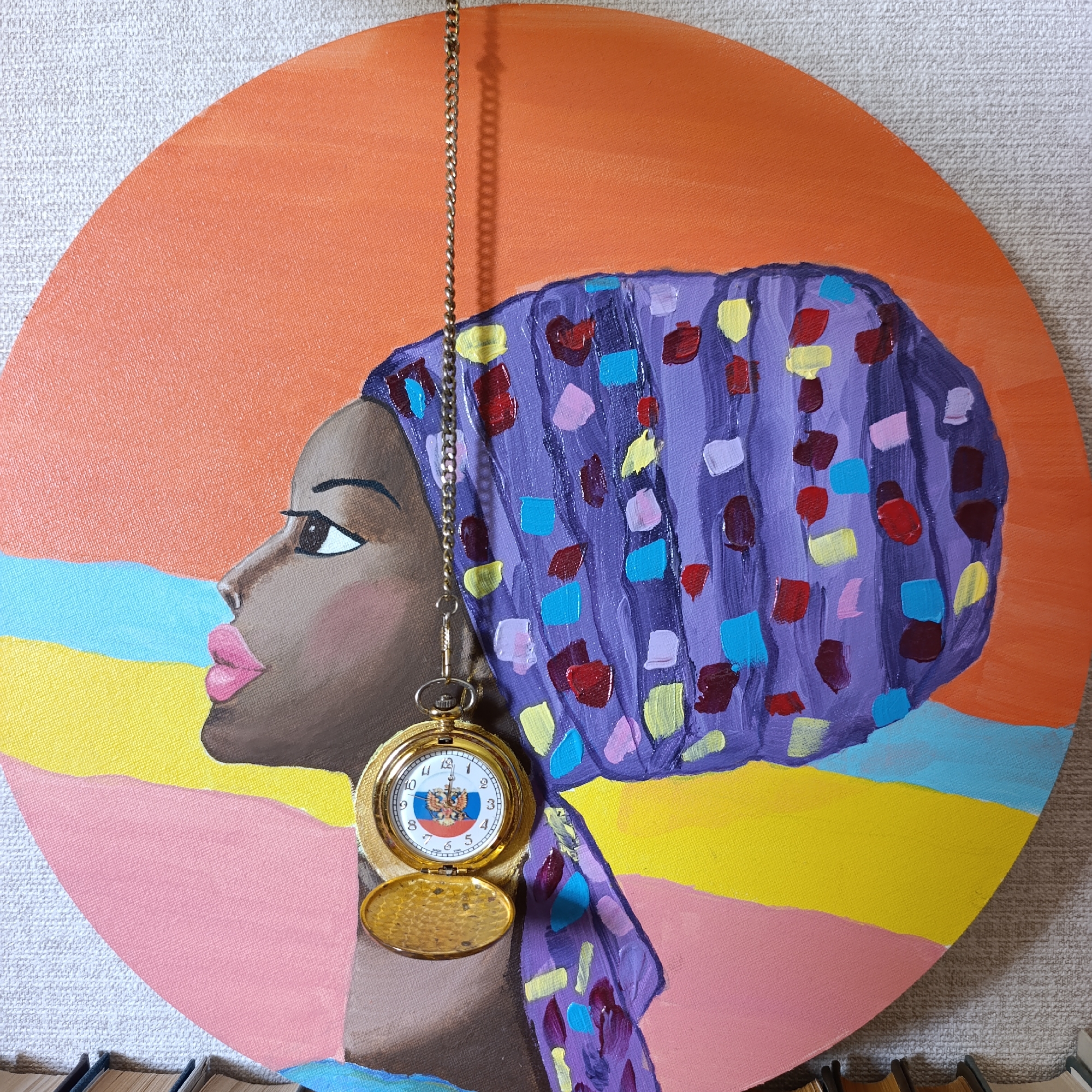 The image depicts a circular acrylic painting on canvas, featuring a woman's profile in a colorful headscarf. The background is divided into horizontal bands of orange, light blue, yellow, and pink. The woman has dark skin, full pink lips, and large brown eyes. She wears a purple headscarf adorned with colorful abstract shapes in red, yellow, light blue, and pink. A gold pocket watch on a silver chain hangs from the headscarf, its cover open, revealing a watch face with Roman numerals and what appears to be the Russian coat of arms in the center. The painting is displayed against a textured, light gray wall. In the foreground, the spines of several books are visible, suggesting the artwork is part of a home or a small display. There is no information in the image to determine the specific location within the city of Марковский, Russia.