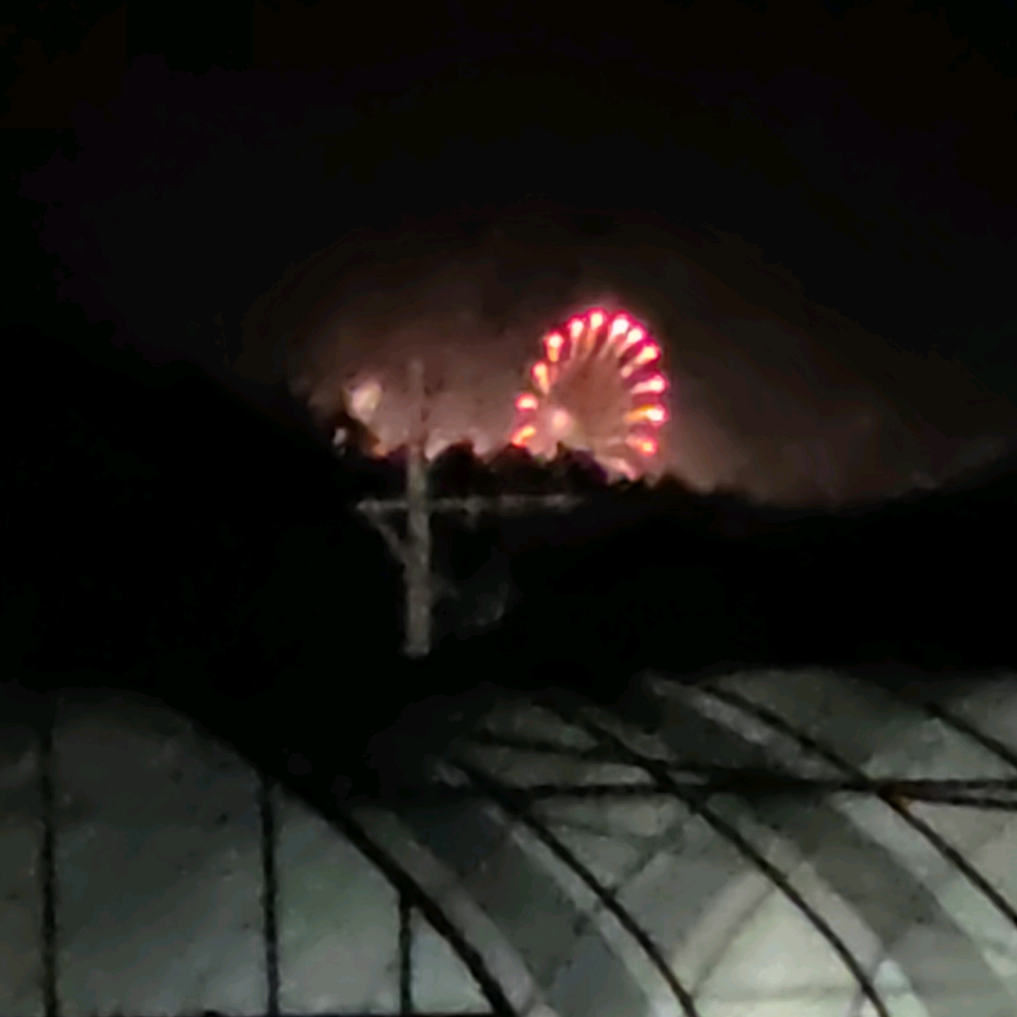 A night time outdoor scene depicts a fireworks display in Dunnae-myeon, South Korea. A Ferris wheel-shaped firework with red-orange lights dominates the upper middle portion of the frame. The firework is set against a dark, hazy sky, likely due to smoke from the display. In the foreground, a dark silhouette of trees or a hill obscures the lower part of the image. A faint, dark, cross-shaped object, possibly a utility pole, is visible in front of the firework. The bottom of the image shows a blurred, light-colored structure, potentially part of a greenhouse or an awning, with visible support beams. The overall image is dark and slightly out of focus.