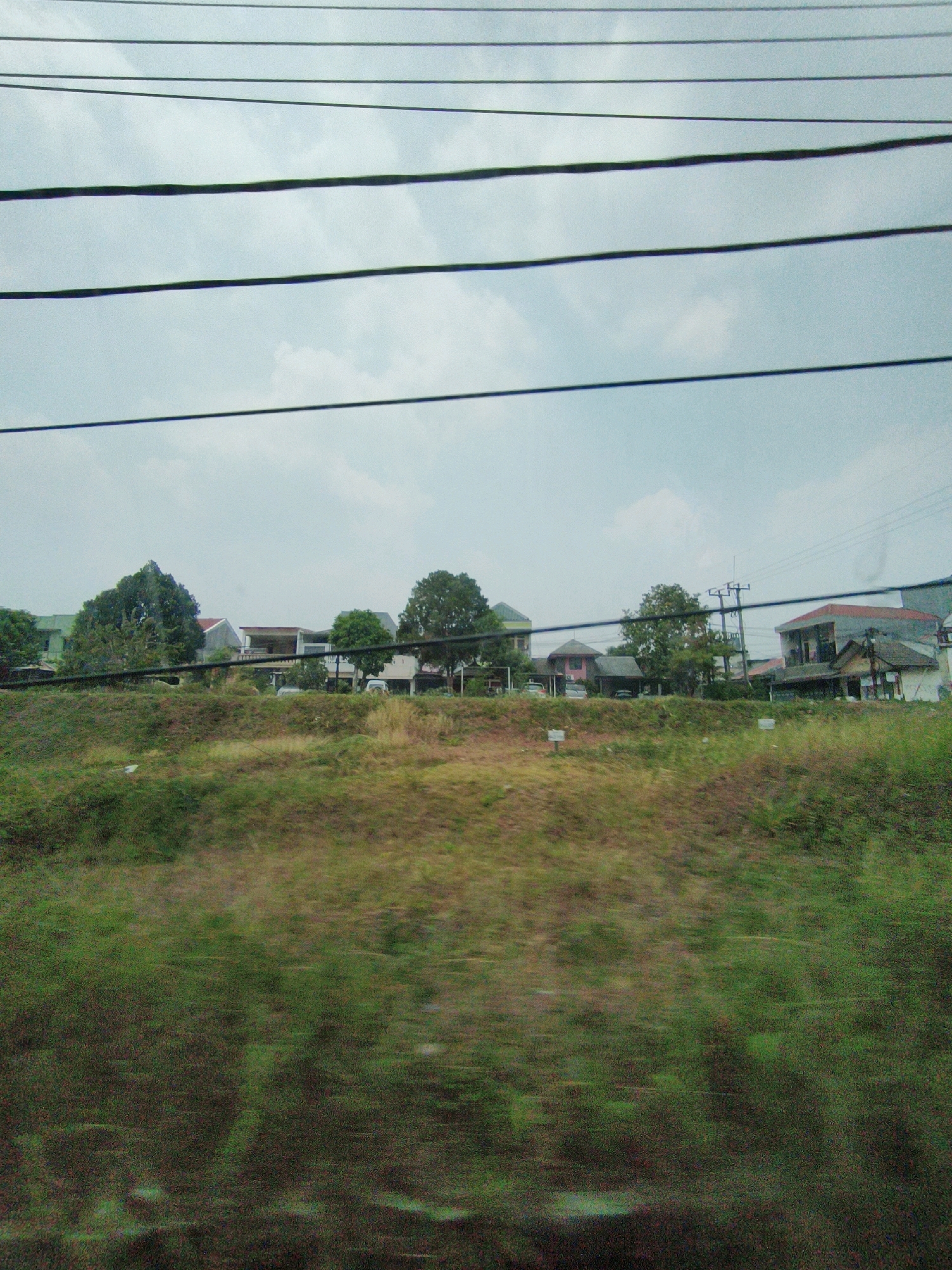 The image depicts a view of a residential area in Sukaraja, Indonesia, seen from a moving vehicle, likely a train or car, as evidenced by the blurred foreground and the presence of overhead wires. In the background, several houses and buildings are visible, nestled among trees. The architecture suggests a mix of modern and traditional styles. A utility pole with wires is prominent in the mid-ground, indicating the presence of electricity. The foreground is a blurred expanse of green vegetation, likely a grassy embankment. The sky is overcast with a light blue hue. No people are clearly discernible in the image. The overall impression is that of a suburban or semi-rural landscape.