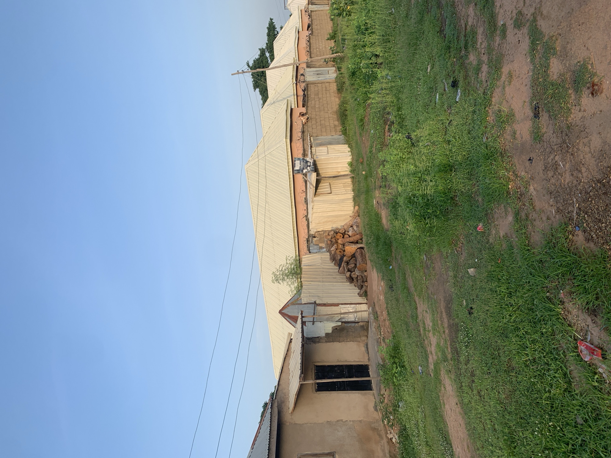 A row of modest, single-story buildings with corrugated metal roofs lines a dirt road in Jalingo, Nigeria. The structures vary in construction, with some appearing to be made of concrete blocks and others of less finished materials like plaster and corrugated metal sheets. Piles of cut logs are stacked against one of the buildings. The area is overgrown with tall grass and scattered debris. Power lines stretch across the clear, pale blue sky. The scene depicts a residential area with a functional, somewhat unkempt appearance.