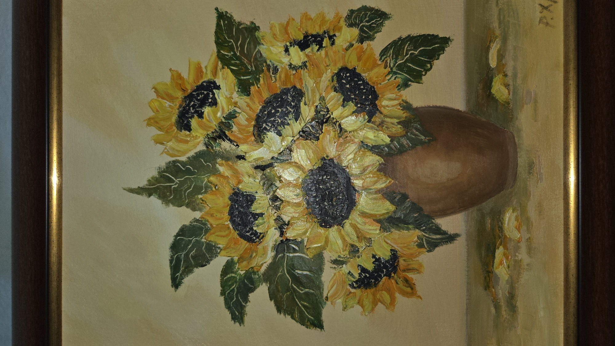 A framed painting depicts a still life of sunflowers in a brown vase. The sunflowers are arranged densely, their yellow petals and dark centers dominating the composition. Green leaves are interspersed among the flowers. The vase is a simple, bulbous shape. The background is a muted beige tone. The painting is framed with a dark brown frame with a gold inner edge. The painting's frame is on top of what appears to be a wall. The location is not discernible from the image.