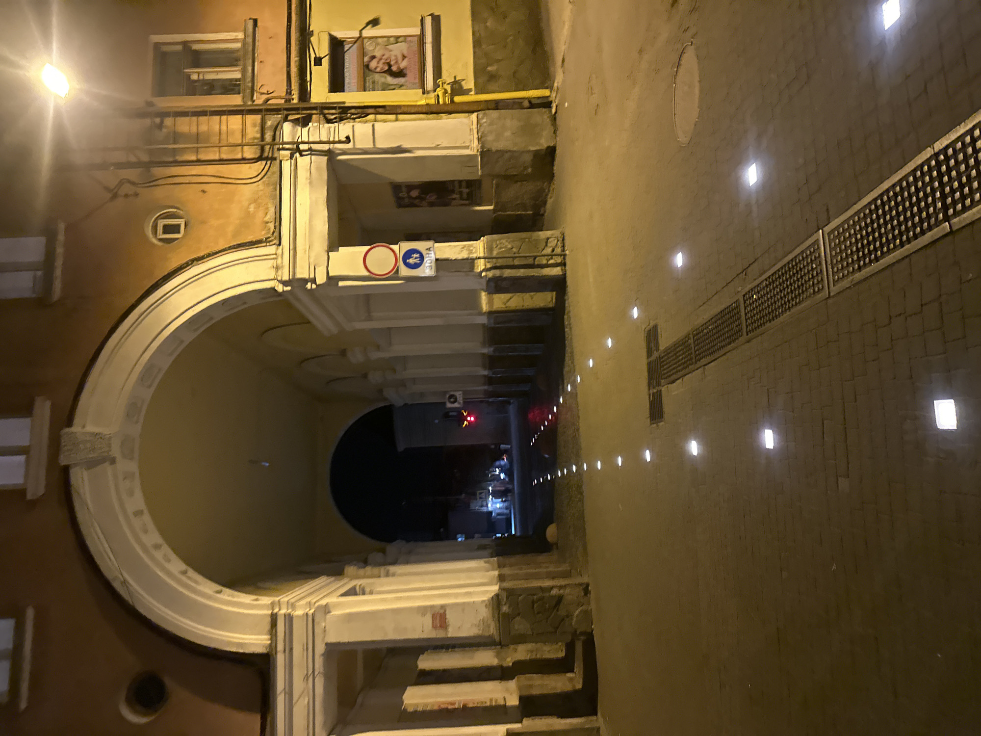This image depicts a nighttime view of an archway and a cobblestone pathway illuminated by embedded square lights and a street lamp. The archway leads to a darkened street with a red traffic light visible in the distance. Signs, including a pedestrian crossing symbol, are present on the right side of the archway. The building facade is a warm, ochre color, with architectural details like pilasters and decorative elements visible. A poster featuring two smiling people is displayed in a window on the right side of the building. A yellow pipe runs vertically along the building's exterior. A metal grate runs along the pathway, parallel to the embedded lights. The overall scene suggests an urban environment at night, possibly in Ternopil, Ukraine, given the context provided.