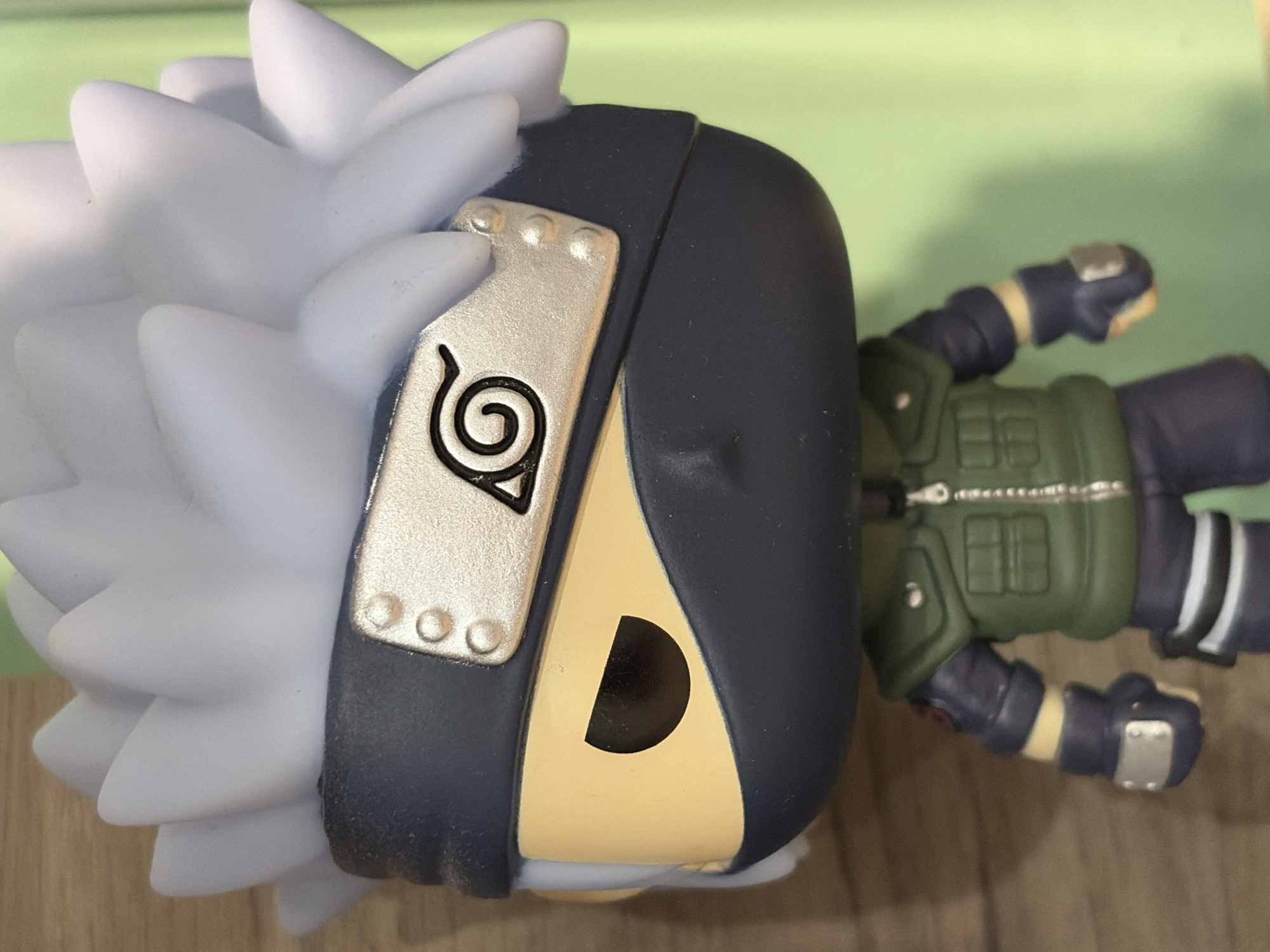 A close-up, eye-level shot shows a Funko Pop figurine of the anime character Kakashi Hatake. The figurine is positioned in the center of the frame, facing forward. It has pale blue spiky hair, a dark blue headband with a silver metal plate featuring the Konoha symbol, and a dark blue mask covering the lower half of its face. The figurine wears a green vest over a dark blue outfit. Its left arm is bent slightly at the elbow, and its left hand is gloved. The background is a blur of warm, light brown wood grain on the left and a solid light green on the right. The lighting is even, highlighting the details of the figurine. The location is not specified in the image.