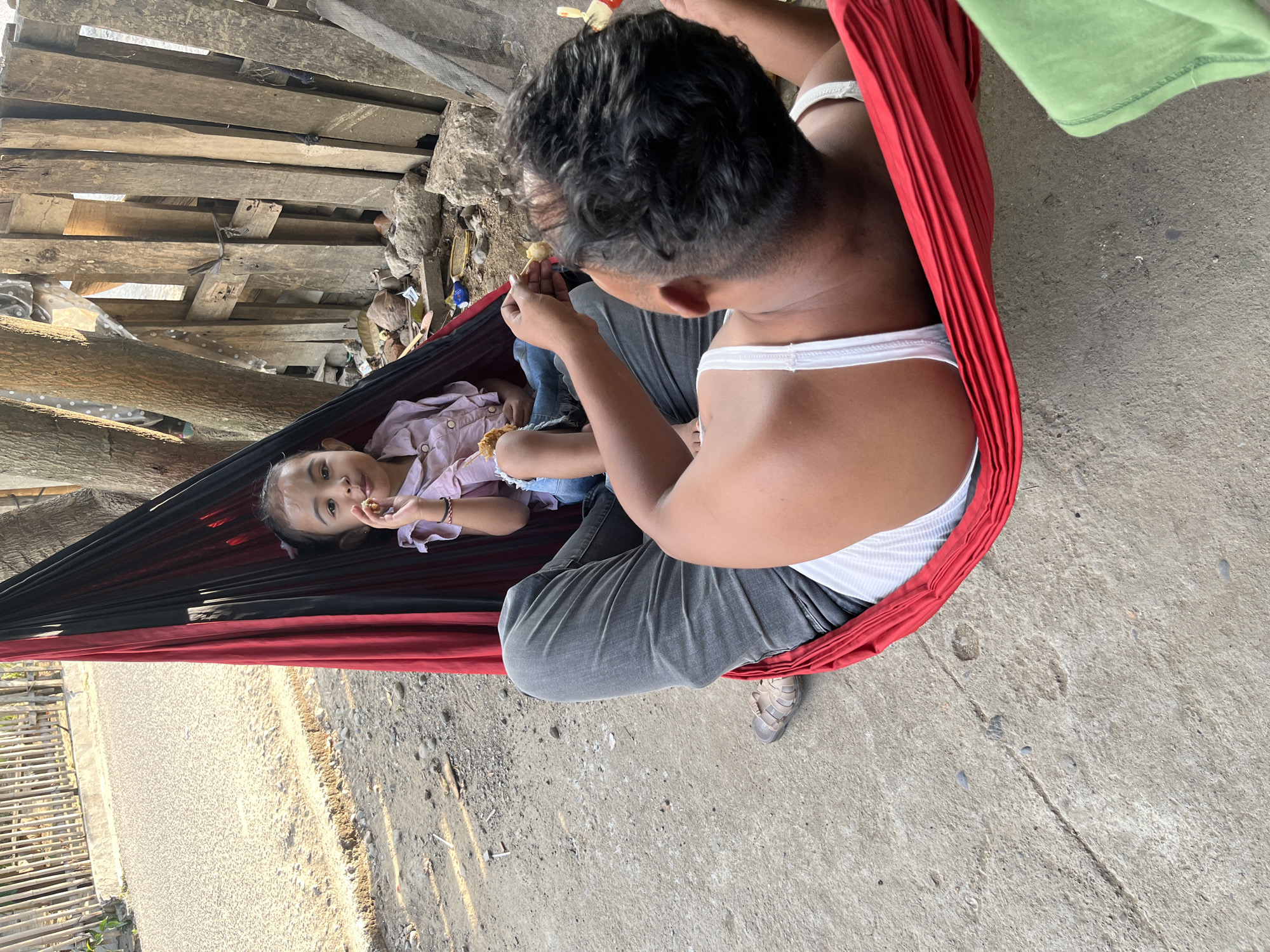 In Kruengmate, Indonesia, a father and daughter are relaxing in a hammock made of red and black fabric. The father, wearing a white tank top and dark grey jeans, is lying down in the hammock, facing away from the camera. His right hand is raised, holding a piece of food. The daughter, positioned above him in the hammock, is wearing a pink collared shirt and blue jeans. She is facing the camera, with a piece of food in her left hand, near her mouth. Both are engaged in eating. The hammock is suspended between two trees, with a rustic wooden fence and scattered debris forming the background. The ground is a mix of concrete and gravel. Sunlight casts shadows, indicating it is daytime.