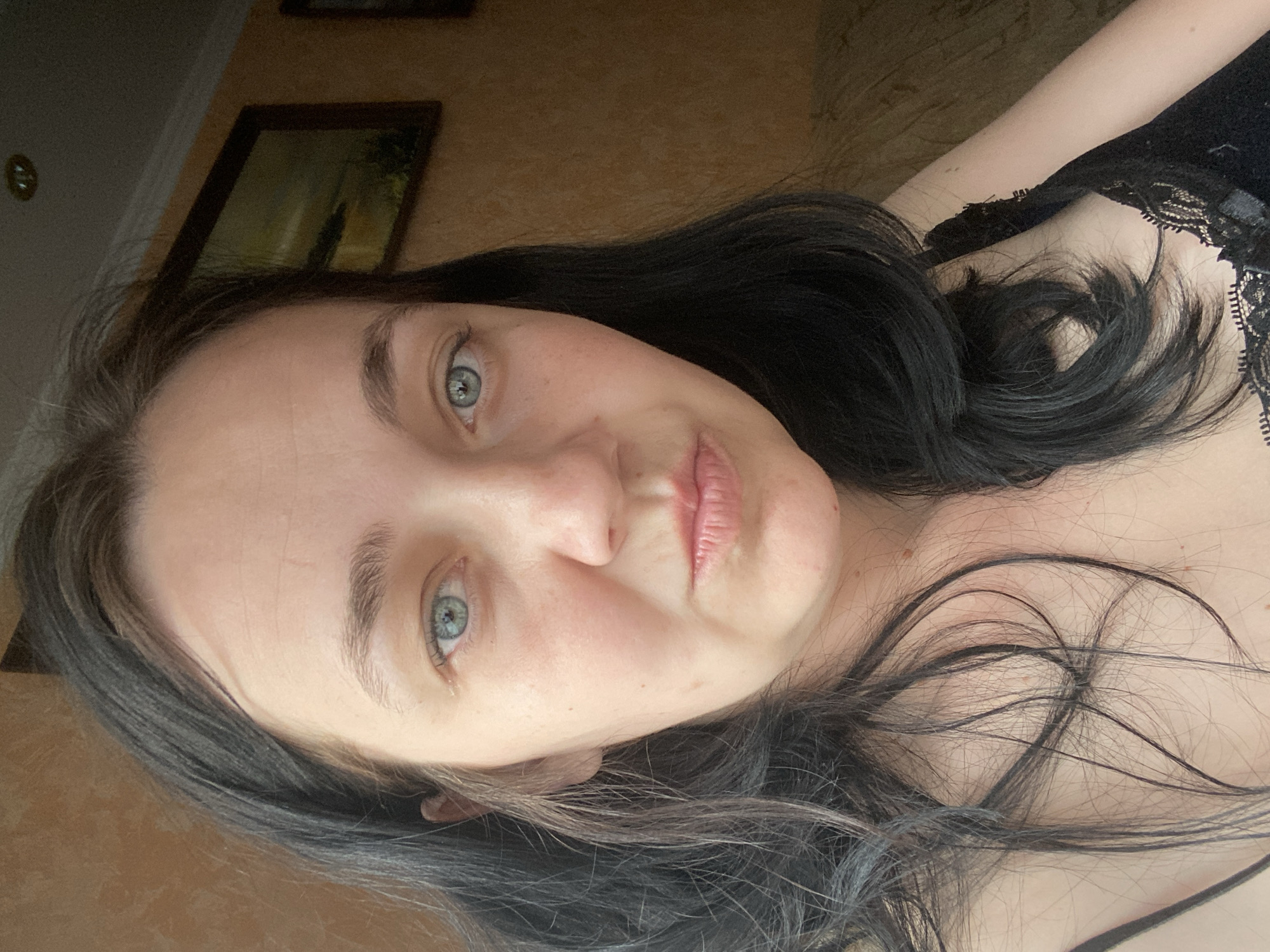 A medium shot portrait of a woman with dark hair and light blue eyes is shown. She is looking directly at the camera with her lips slightly pursed. The background is out of focus and appears to be an indoor setting with textured walls and a framed picture on the wall. The woman is wearing a black lace camisole.