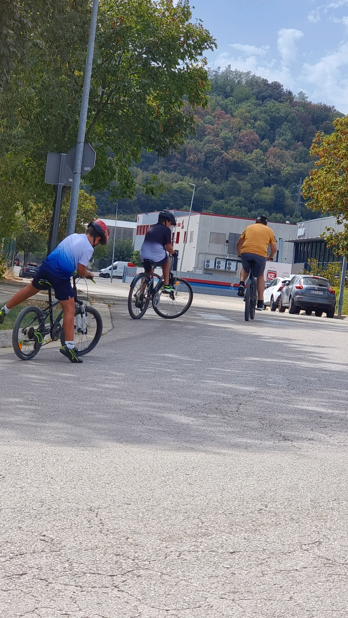 Three individuals are cycling on a paved road. In the foreground, a person wearing a blue and white t-shirt, dark shorts, and a red helmet is riding a black bicycle with green accents on the wheels. Their left leg is extended backward. To the left of this cyclist, a grey lamppost with a sign attached is visible. Further down the road, a second person wearing a dark t-shirt and shorts, and a black helmet, is riding a dark bicycle. In the background, a third person wearing a bright orange t-shirt and dark shorts is riding a bicycle. Behind the cyclists, there are several parked cars, including a grey SUV with license plate "8023 GYY". A modern building with windows and a prominent red sign is situated behind the cars, and further in the distance, a tree-covered hill rises under a cloudy sky. The overall setting appears to be an urban or industrial area, possibly on the outskirts of Olot, Spain, given the architecture and natural landscape.
