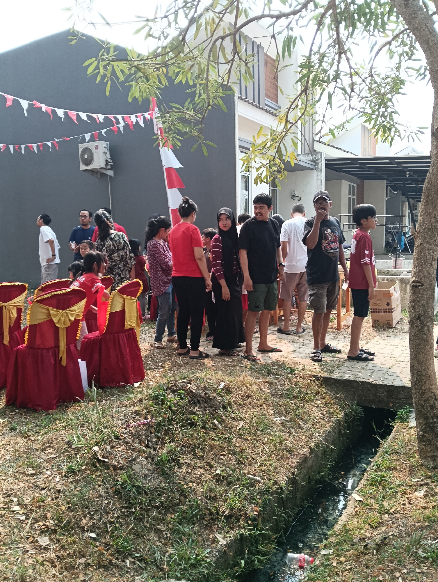 In Parung Panjang, Indonesia, an outdoor gathering is underway near a residential area. A group of approximately 15 individuals, varying in age and gender, are present. Some are standing in groups, while others are positioned individually. Decorative red and gold chairs are set up, covered with draped red fabric and gold sashes. Several Indonesian flags, red and white, are visible, with one unfurled and attached to a pole and others strung as a garland across a building. The environment includes a grey building with an air conditioning unit mounted on its exterior wall, and a narrow, overgrown drainage ditch running alongside a paved pathway. The ground is covered with dry leaves and sparse vegetation. The lighting suggests it is daytime, possibly late morning or early afternoon, with shadows indicating the sun's position.
