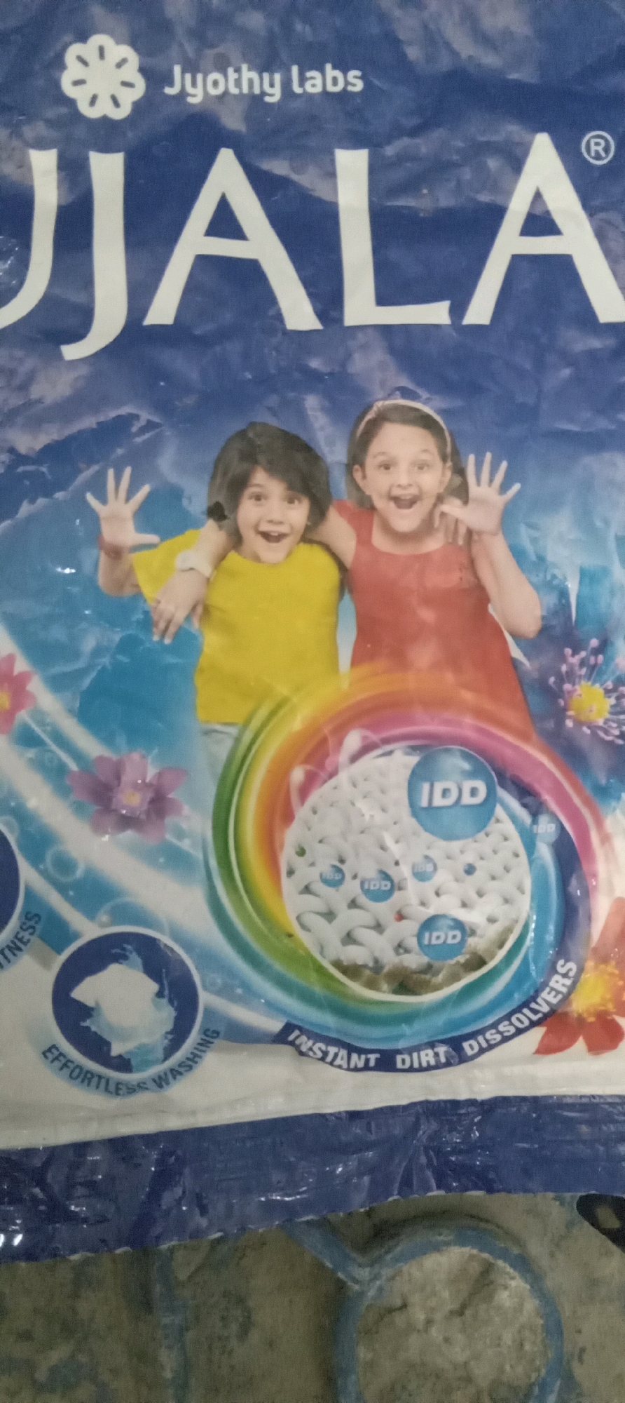 This image shows a close-up of a detergent packaging, likely for "Ujala" brand, featuring a floral logo and "Jyothy Labs" text at the top. Two young girls are depicted on the packaging, both smiling and appearing to wave. One is wearing a yellow t-shirt, and the other a red dress, and they are positioned in front of a blue, watery background with floral elements. A circular graphic in the center of the packaging shows a magnified view of detergent granules, some labeled with "IDD," and surrounded by a rainbow-like swirl. Text on the packaging includes "EFFORTLESS WASHING" and "INSTANT DIRT DISSOLVERS." The packaging itself is slightly creased and appears to be made of plastic. The background is a mottled, textured surface, possibly a floor or a wall, with a blue plastic handle or edge visible at the bottom. There is no indication of the specific location within Vaddahalli, India, beyond the context of the product packaging itself, which implies its distribution or origin in India.