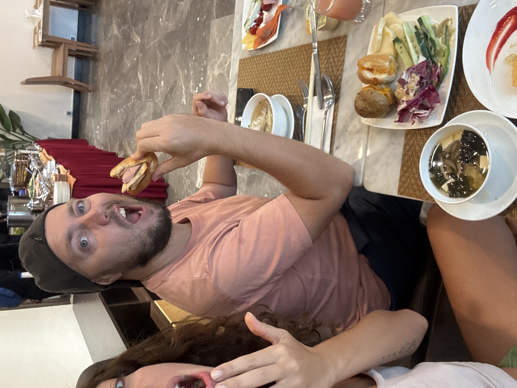 A man and woman are seated at a table in what appears to be a dining area. The man is wearing a gray hat and a pink shirt, holding a small sandwich up to his mouth with an exaggerated expression. The woman is wearing a white shirt. On the table are several plates of food, including salads, fruit, and soup. There is also silverware and a glass of juice. The location may be a hotel or restaurant in Đà Nẵng, Vietnam, given the style of the room and the food on the table.