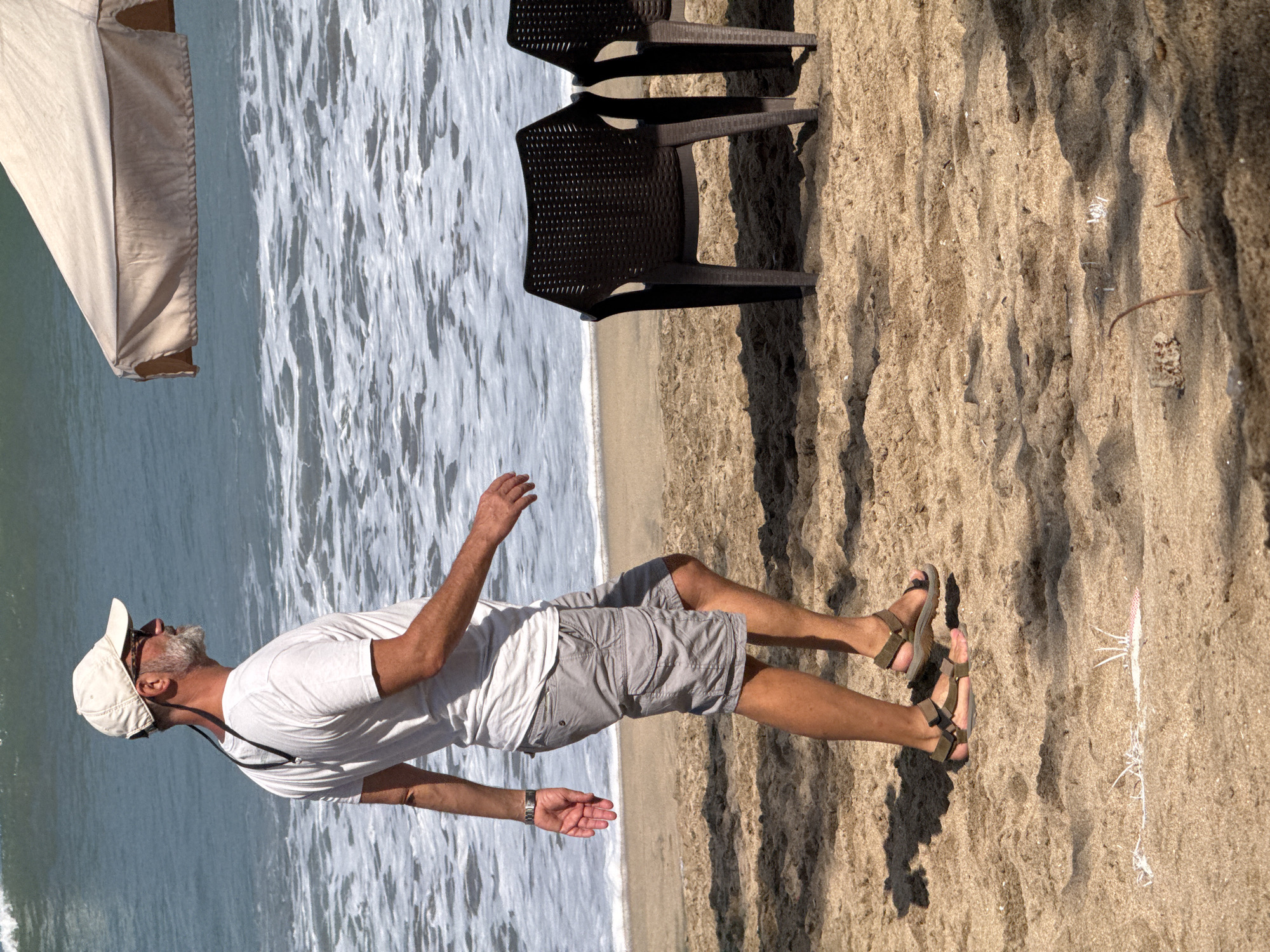 An adult male is walking on a sandy beach near the ocean in Kuta, Indonesia. He is wearing a white t-shirt, grey cargo shorts, a white baseball cap, sunglasses, and brown sandals. He has a beard and is carrying a lanyard around his neck. The ocean is visible in the background with waves breaking on the shore. Two dark plastic beach chairs and a light-colored umbrella are also present on the sand. The sun is casting shadows on the sand.