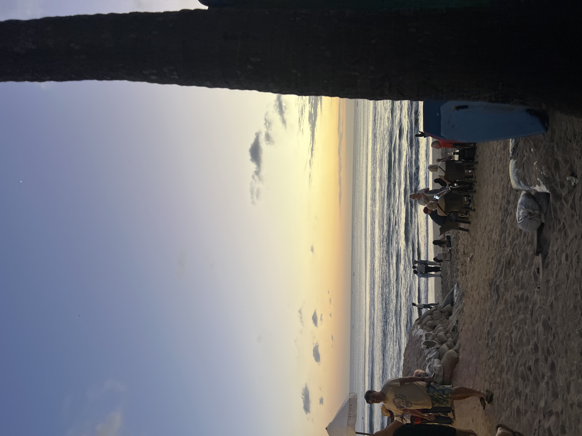 A vertical, medium shot captures a beach scene in Kuta, Indonesia, during twilight. The sky transitions from a pale blue at the top to a soft yellow and orange near the horizon, where the ocean meets it. Gentle waves roll onto the sandy shore.
In the foreground, a large, dark tree trunk is on the right side of the frame. The sand is littered with a few scattered white bags and a blue boogie board propped upright. A group of people are visible in the middle ground, some sitting at tables and chairs set up on the sand, appearing to be at an outdoor cafe or restaurant. Others stand and converse. A white umbrella stands near the left edge of the frame.
Further back, near the water's edge, a few more individuals are present. The overall atmosphere is relaxed, with people enjoying the evening on the beach. The scene is dimly lit, characteristic of sunset or dusk.