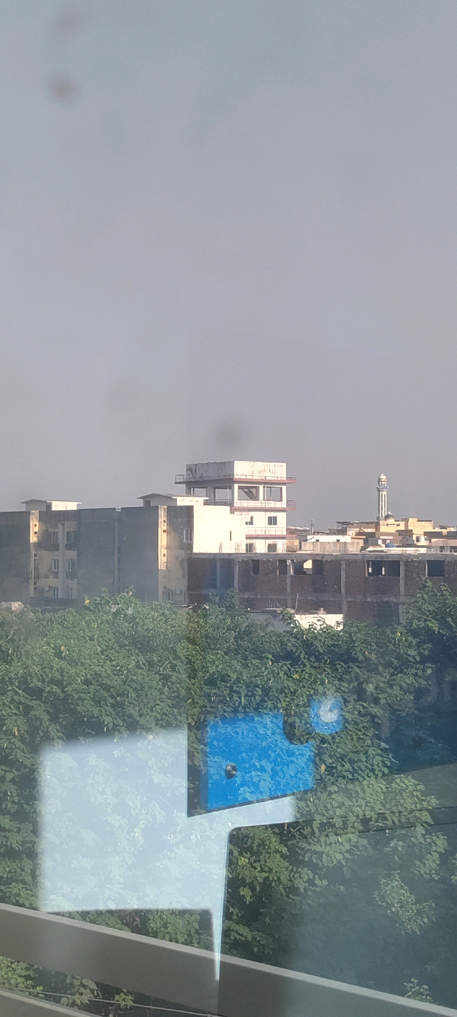 A hazy, overcast sky dominates the upper portion of the image. Below, a cluster of buildings, likely part of the city of Khana Dak, Pakistan, is visible. The structures include a tall, modern white building with multiple levels and balconies, and adjacent to it, a darker, more industrial-looking building with visible construction elements. Further to the right, a minaret of a mosque pierces the sky, indicating the presence of a religious site within the urban landscape. In the foreground, dense green foliage obscures the lower part of the buildings, suggesting a lush, tree-lined area. Reflections on a glass surface in the very bottom of the image show abstract shapes of light blue and white, overlaying the scene. There is a plume of dark smoke visible rising from behind the white building, indicating a potential fire or industrial activity. No people are directly visible in the frame.