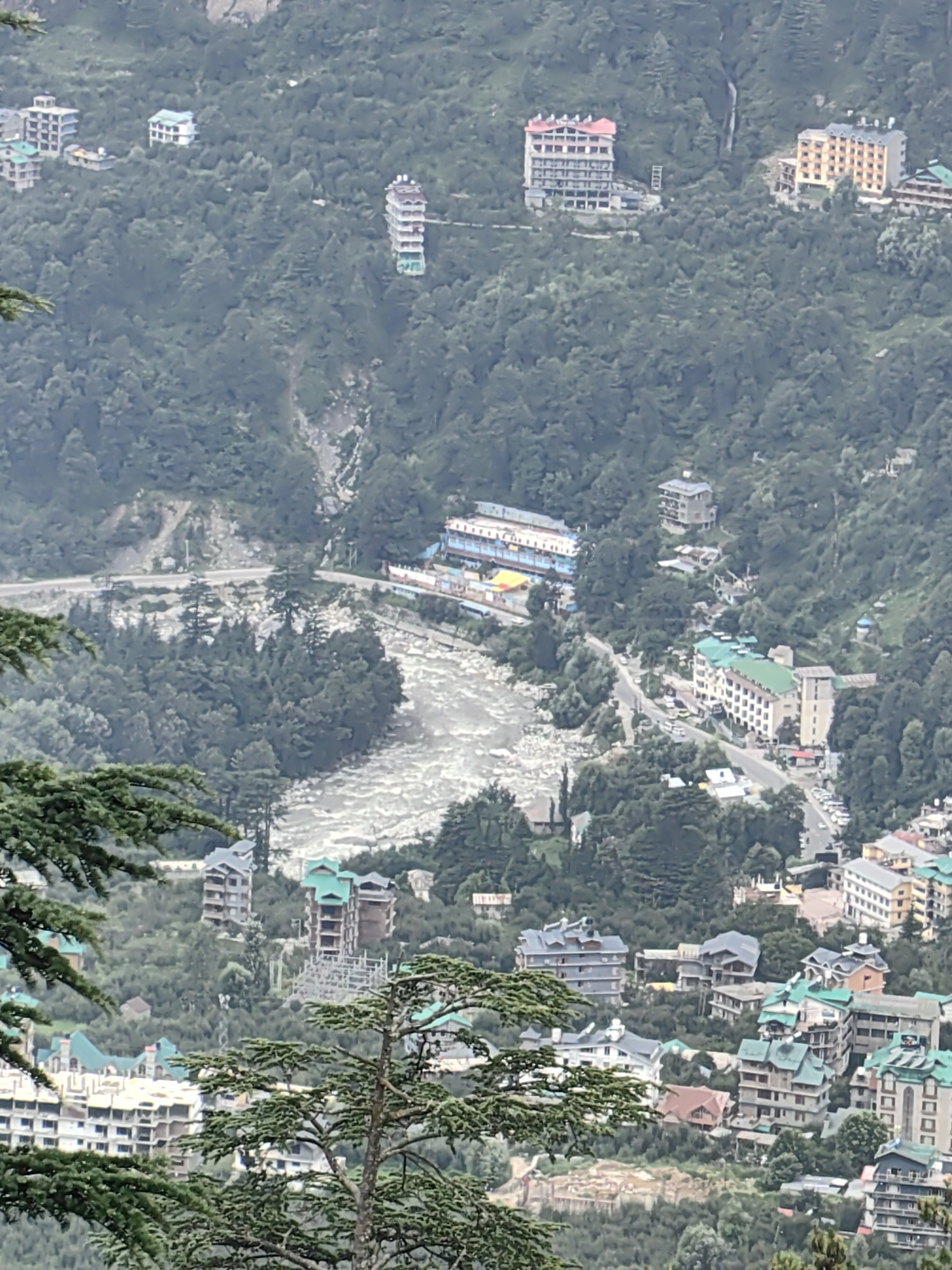 An aerial, high-angle shot shows a hazy, overcast day in Prini, India, a town nestled in a lush, mountainous region. A wide, turbulent river flows through the valley, flanked by dense, dark green trees. Buildings of various sizes and architectural styles are scattered across the slopes. Some appear to be hotels or resorts, with multiple stories and flat or sloped roofs, some with green tiling. Several structures are under construction, with exposed frameworks visible. A road winds through the town, with a few cars parked along its side. The overall impression is of a developed hillside settlement within a natural, forested environment.