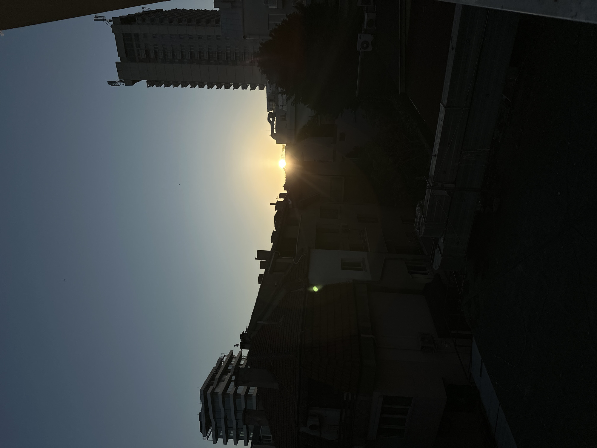 The image shows a cityscape at sunset in Varna, Bulgaria. The sun is positioned between two buildings, creating a bright glow with sun rays emanating from it. On the left of the sun, a modern high-rise apartment building is visible. On the right, an older building with an orange-red roof is visible, accompanied by trees and other lower buildings. The sky is clear with a gradient of blue.