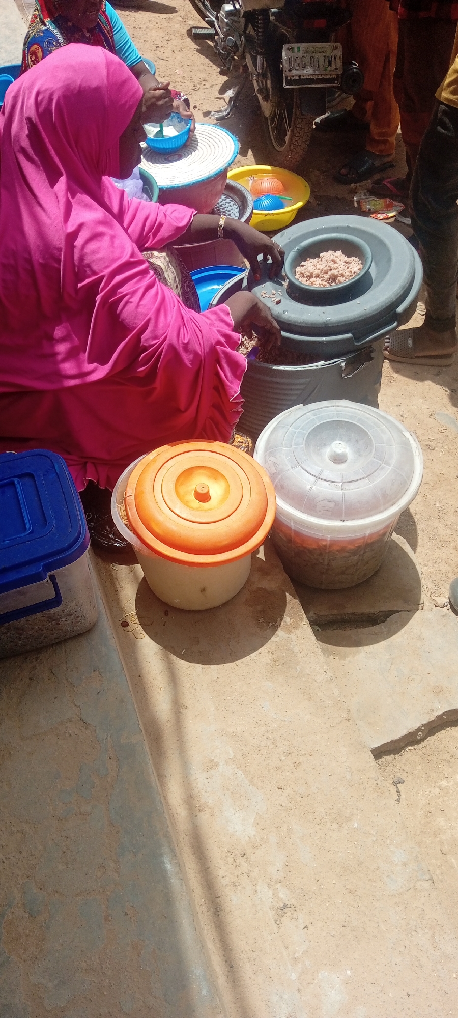 The image depicts a scene outdoors, likely in a dusty, sun-drenched environment with a light-colored, rough ground surface. The time of day appears to be daytime, with harsh shadows cast by the sun.
The central subjects are several individuals engaged in preparing food. A person wearing a bright pink hijab and matching garment is actively sorting through a large container of small, grain-like food items. Their hands are visible, manipulating the food. To their left, another person, partially visible, is also involved in food preparation, holding a small blue bowl.
Various containers are present, indicating a food preparation or distribution setting. There's a large gray container with a smaller bowl of food on top, likely where the main sorting is happening. Below this, a large cylindrical container is visible. To the left of the pink-clad individual is a blue plastic container with a lid, also appearing to hold food items. In the foreground, there are two lidded buckets: one with a bright orange lid and a pale body, and another with a translucent white lid, revealing a mixture of food inside. In the background, a motorcycle is parked, with a license plate visible that reads "UGG 017WY FOR REGISTRATION". There are also other people in the background, their attire suggesting a warm climate.
The scene appears to be a communal food preparation activity, possibly for a meal or distribution. The overall atmosphere is one of industrious activity in a basic, outdoor setting. The visible text on the motorcycle license plate provides a specific detail about the location context.