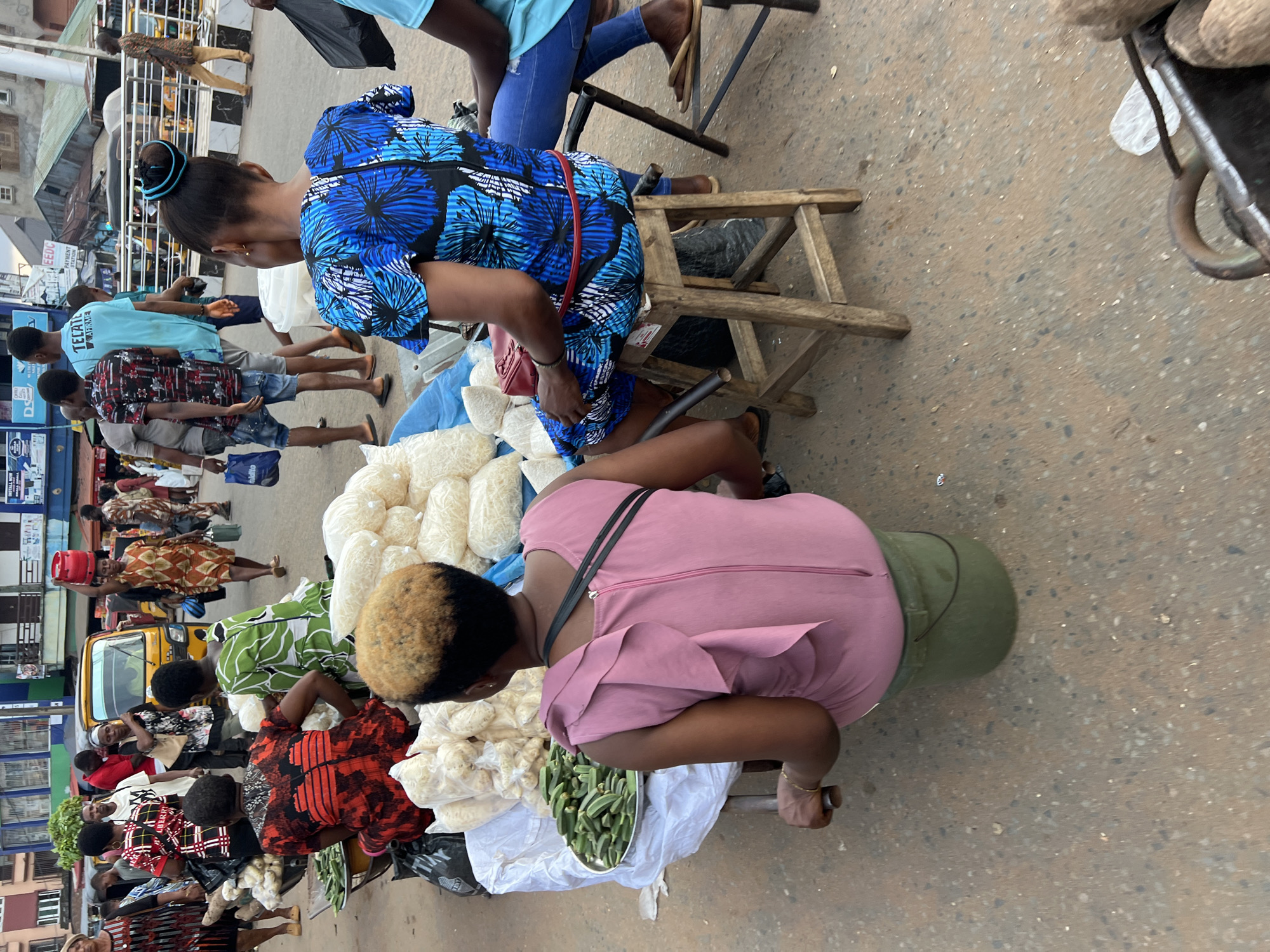 A street market scene in Nkpor, Nigeria, captures individuals engaged in commerce. In the foreground, two women are positioned behind stalls laden with produce. The woman on the left, wearing a pink dress, is seated on a green bucket and appears to be tending to her goods. To her right, another woman in a vibrant blue and black floral dress is seated on a wooden stool. Behind them, a variety of food items, including what appears to be yams and okra, are displayed. The market is bustling with other people, some carrying items and others interacting. A yellow auto-rickshaw, commonly known as a 'Keke Napep,' is visible in the background, suggesting transportation is readily available. The environment appears to be a paved street with buildings and shops lining the sides, indicating a commercial district within the city. The overall atmosphere is one of daily life and economic activity.