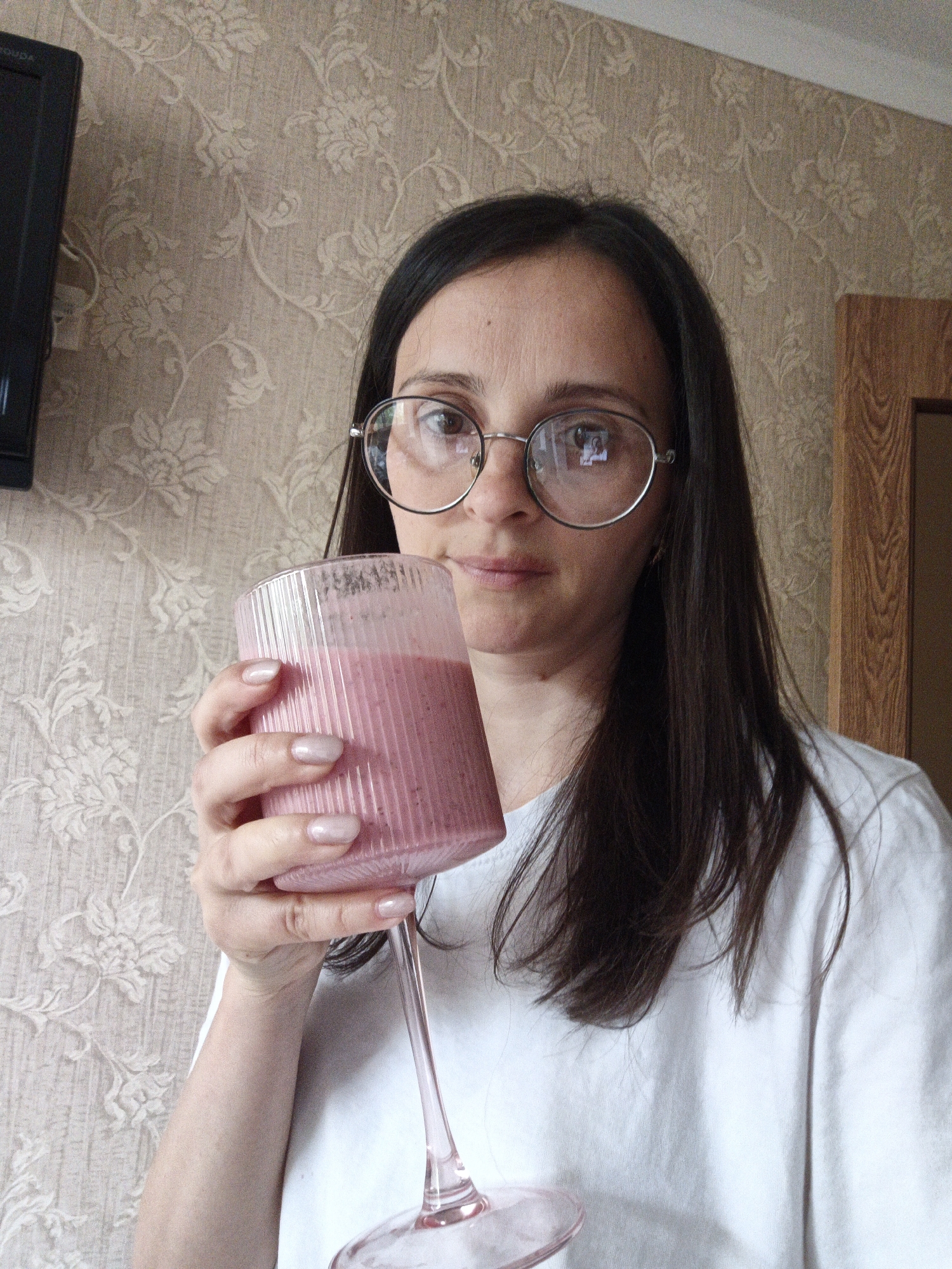 A woman with dark hair and round glasses is holding a glass of pink smoothie in front of a patterned wallpaper. The room contains a dark TV on the wall and a wooden door frame. The location is Goryachiy Klyuch, Russia.