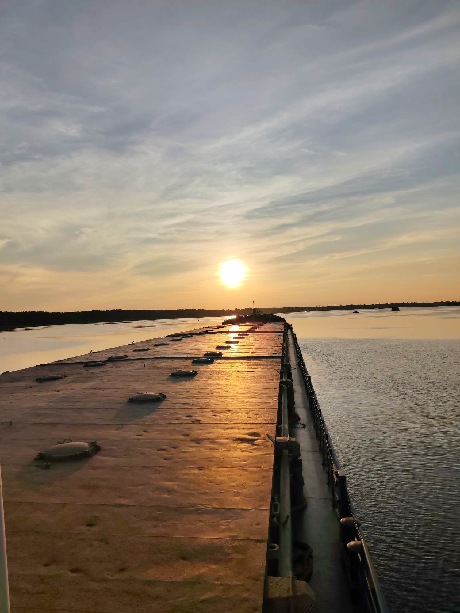 A low-angle, wide shot captures a serene sunset over a body of water, with a long, flat structure dominating the foreground. This structure, possibly a barge or pier, is covered in a weathered, tan material, punctuated by numerous circular, raised hatches. The surface is illuminated by the warm, golden light of the setting sun, which is positioned low in the sky, just above the horizon. Wispy clouds streak across the sky, reflecting the sunset's colors. The water is calm, with gentle ripples catching the light. In the distance, a dark, tree-lined shore is visible, and a few small objects, possibly buoys or distant boats, dot the water. The scene is devoid of people. The location is identified as Shchetinino, Russia.