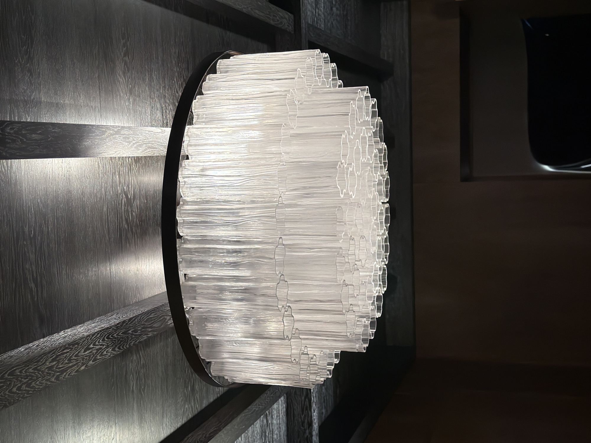 An interior shot taken from below shows a modern, tiered chandelier with clear glass rods suspended from a dark wooden beamed ceiling. The chandelier has a dark circular frame and is illuminated, casting light upwards onto the ceiling. The ceiling features a wood-paneled design with visible beams and a dark, textured finish. The lower portion of the image shows a dark wall and the edge of what appears to be a dark, curved object, possibly a piece of furniture. The scene is dimly lit, with the chandelier being the primary light source. The location is a dimly lit interior space, possibly a restaurant or a high-end residential property, in Moscow, Russia.