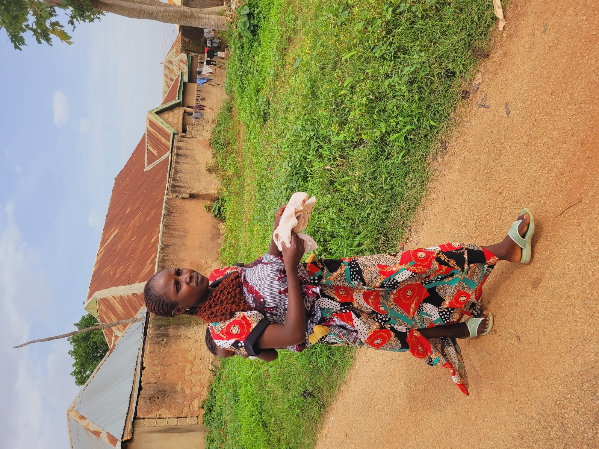 In Bukuru, Nigeria, a woman with braided hair carries a baby on her back. She is wearing a colorful, patterned dress and light-colored sandals. She holds a light-colored cloth in her hands. The woman walks on a dirt path, with lush green grass bordering the path. In the background, several buildings with corrugated metal roofs are visible, along with laundry hanging on a line. The sky is partly cloudy.