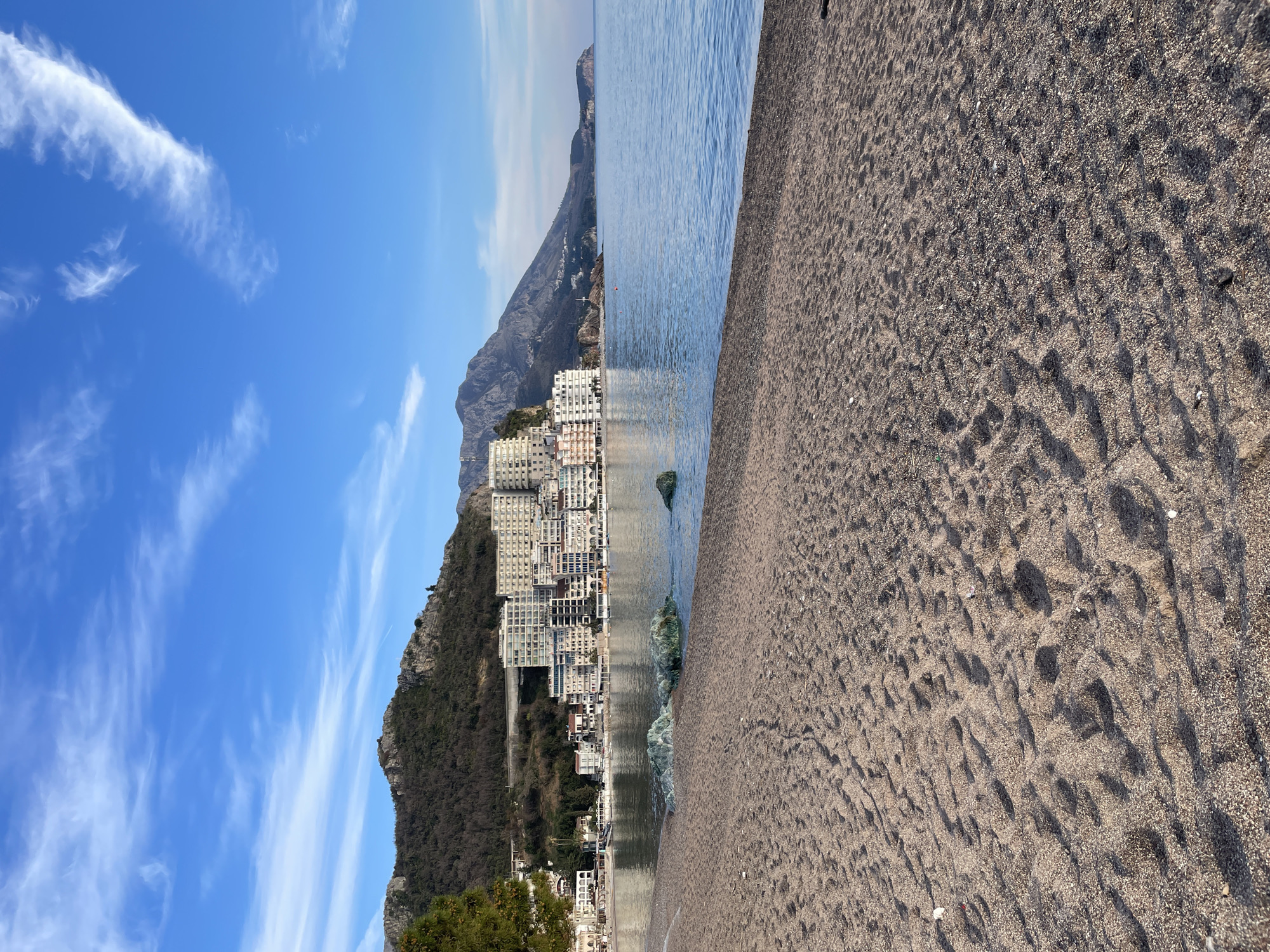 The image displays a panoramic view of the beach and coastal town of Rafailovići, Montenegro, under a clear blue sky with scattered cirrus clouds. In the foreground, a wide expanse of light brown, pebbly sand beach stretches across the lower half of the frame, showing numerous footprints and minor debris. The midground features calm, blue-green seawater, with several partially submerged, irregular rocks exhibiting green coloration near the shoreline. Further out in the water, a few small red buoys are visible. On the opposite side of the bay, the town of Rafailovići is visible, characterized by a dense cluster of multi-story buildings, predominantly white and light-colored, constructed along the waterfront and ascending the lower slopes of a mountain. These buildings often feature multiple balconies. The background consists of a large, rugged mountain covered with sparse, dry-looking vegetation, rising directly behind the town. To the right, the coastline extends into the distance, revealing additional landmasses and a distant headland. No individuals or overt activities are present in the scene.