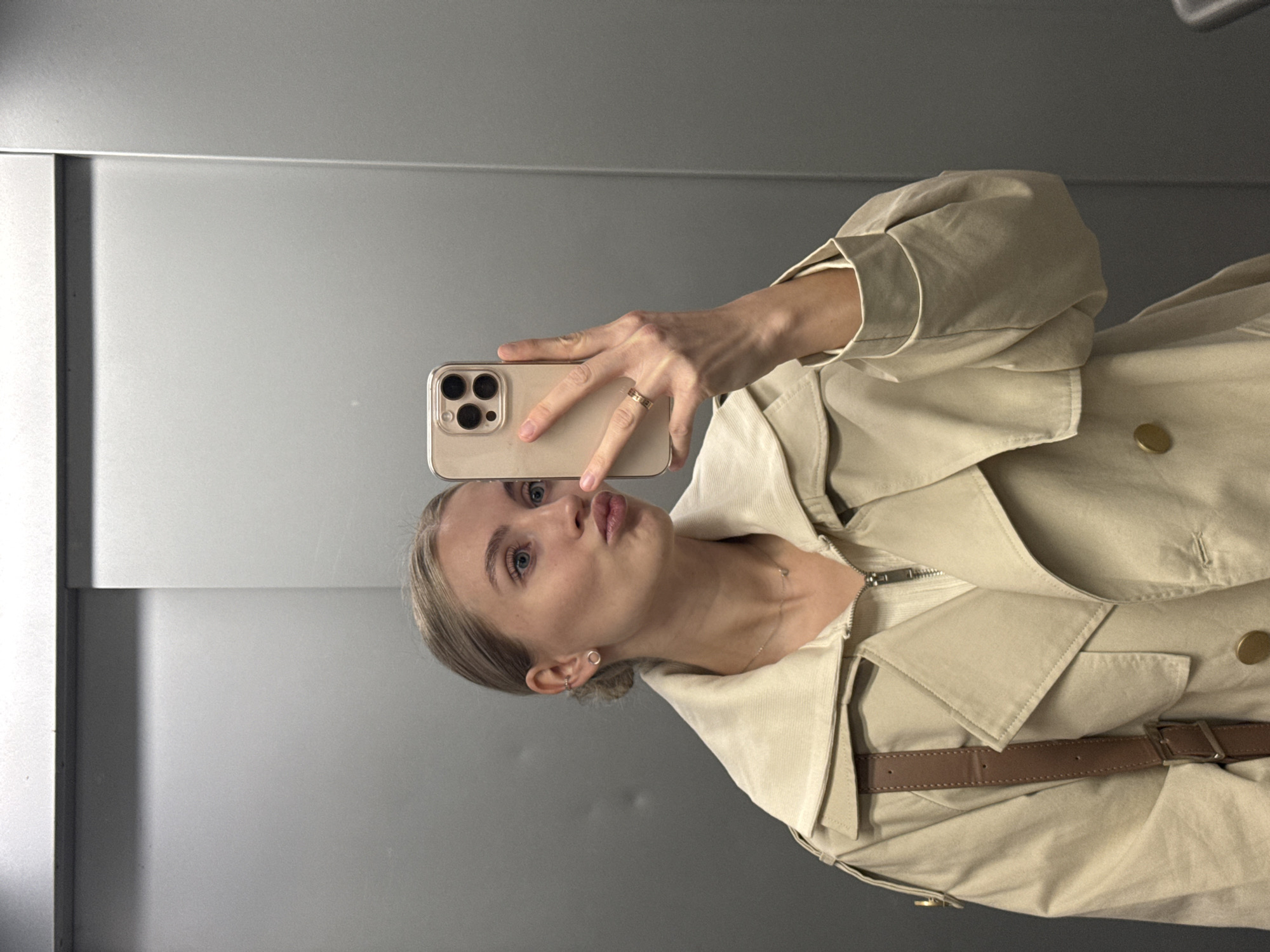 A young, fair-skinned woman with blonde hair pulled back in a bun is taking a selfie in front of a light gray wall, possibly a mirror. She is wearing a beige trench coat with gold buttons and a brown leather belt. She is holding a beige-colored smartphone up in her right hand, which is adorned with a gold ring. Her lips are pursed in a pouting expression. The location is not evident in the image and cannot be identified as Izhevsk, Russia, or any other city.