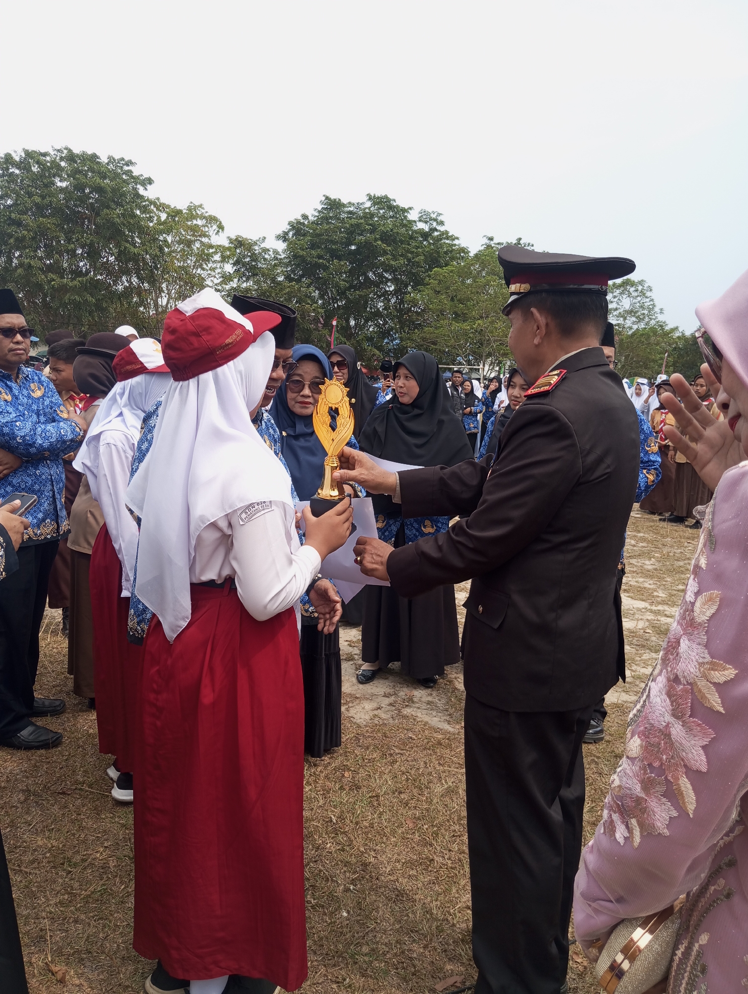 In Pematang Reba, Indonesia, a man in a dark brown suit and cap, possibly a military or police officer, is presenting a gold trophy to a young girl wearing a white hijab and a red skirt. The girl is also wearing a red and white cap. She is holding a piece of paper and appears to be receiving the award. Several other people, including men and women in traditional Indonesian attire and some in school uniforms, are watching the presentation. The scene takes place outdoors on a dry, grassy field, with trees visible in the background. The sky is overcast.