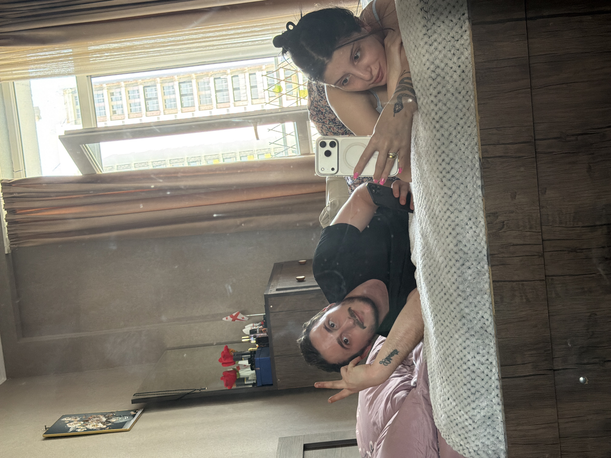A man and a woman are in a bedroom, taking a selfie in a mirror. The man is lying on his side on a bed, wearing a black t-shirt, and holding up two fingers in a peace sign. He has a tattoo on his left arm. The woman is lying on her stomach next to him, also on the bed, wearing a floral-patterned top and holding a smartphone. She has tattoos on her arms. The room has a wooden headboard, a textured white blanket, and pink pillows. In the background, there is a wooden dresser with some items on top, including red flowers and a small Georgian flag. A window is open, revealing a cityscape with buildings, suggesting the location is in Tbilisi, Georgia. The overall impression is a casual, domestic scene.