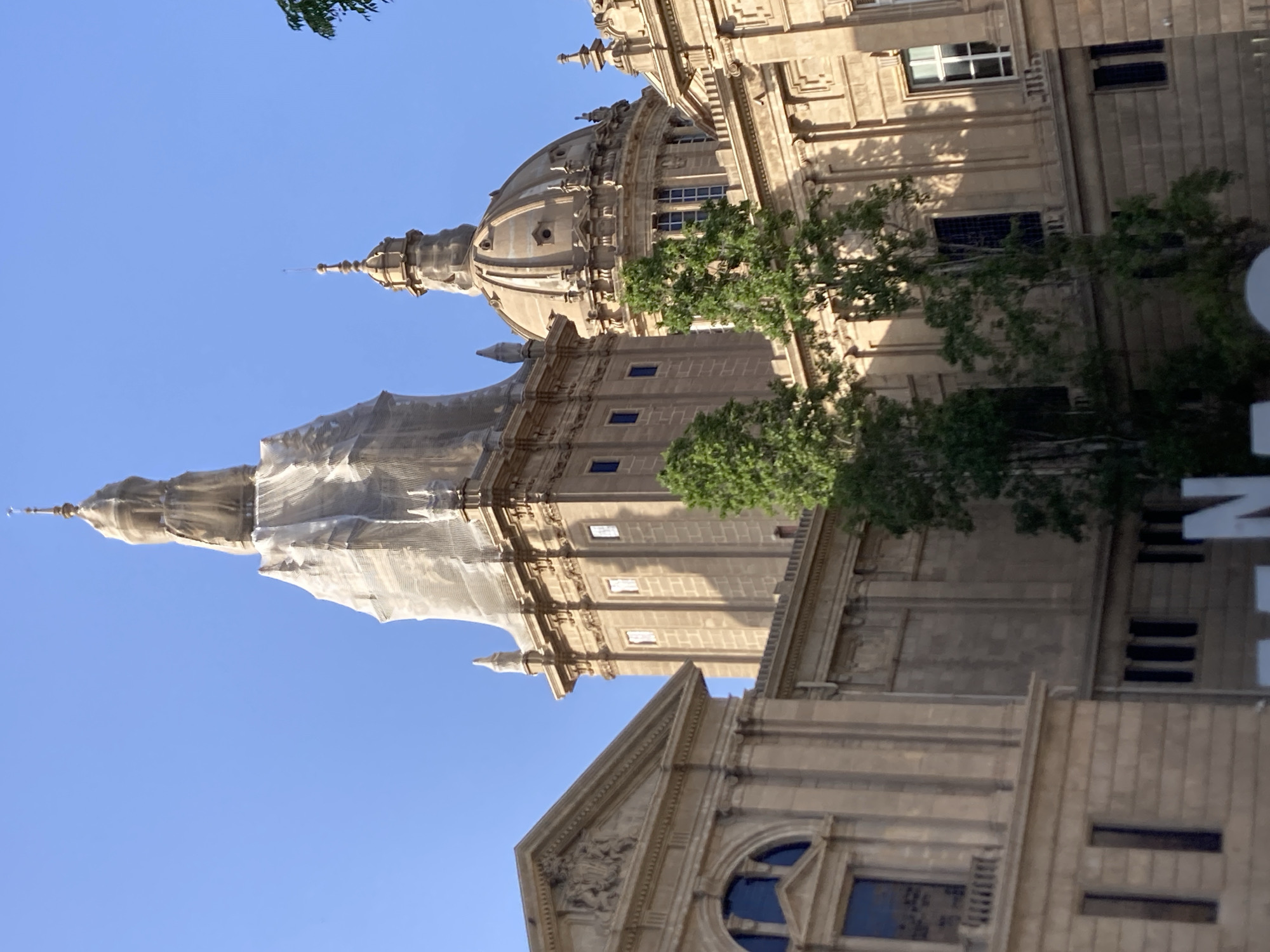 The image shows the Palau Nacional in Barcelona, Spain. The building's exterior is made of stone and features classical architectural elements. Two large domes are visible, one of which appears to be under construction or renovation, covered in netting. The sky is clear and blue. In the foreground, several trees with green foliage are present. The lighting suggests it is daytime, possibly late afternoon due to the shadows cast on the building. There are no people visible in the image.