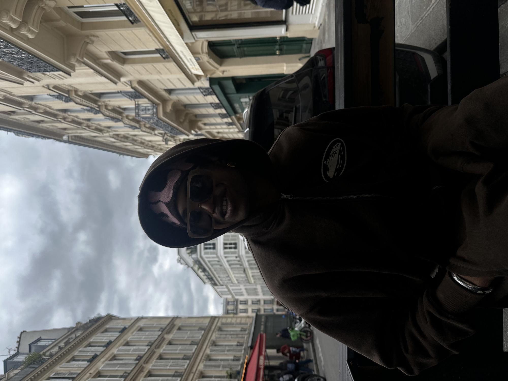 A person wearing a brown hooded sweatshirt and sunglasses is sitting at an outdoor cafe or restaurant in Paris, France. Tall, classic Parisian buildings line the street in the background, and the sky is overcast. Motorbikes are parked along the curb.