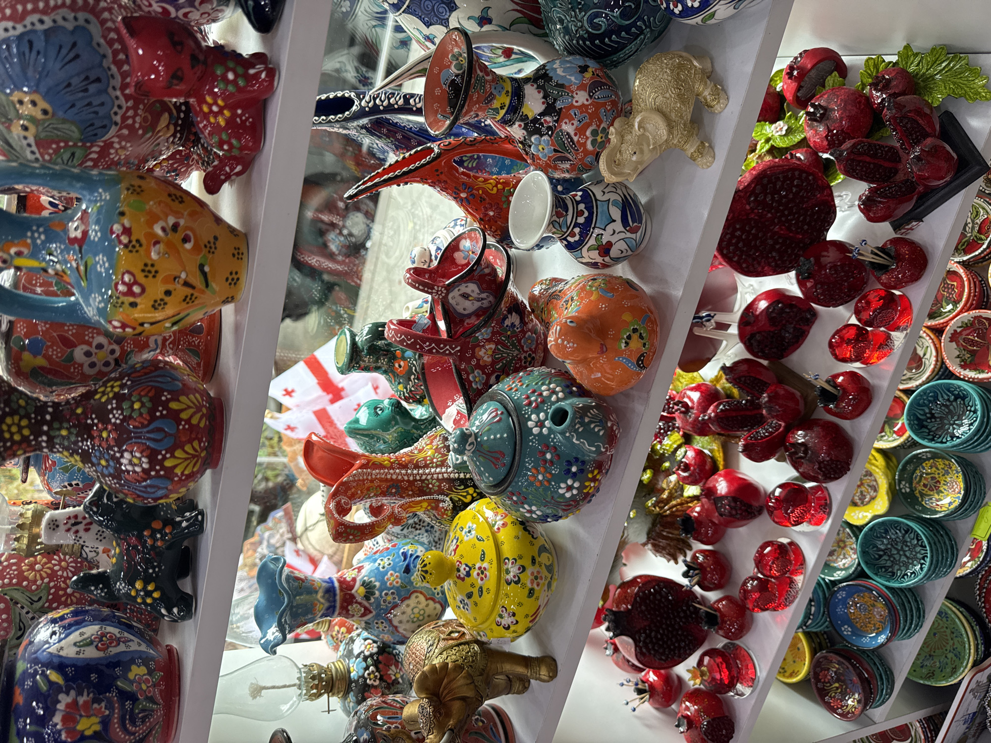 The image depicts a vibrant display of decorative ceramic items on multiple shelves, likely in a shop or market stall in Batumi, Georgia. The shelves are filled with an assortment of colorful and intricately painted vases, bowls, figurines, and decorative objects.
On the top shelf, there are large, ornate vases with floral patterns in blues, reds, and yellows. Smaller figurines, including a black dog and a red cat, are placed between the larger items. Several other ceramic pieces with intricate designs are visible on this shelf.
The middle shelf showcases more decorative vases and vessels, some with animal motifs like an elephant. There are also unique pieces like a red basket with a handle and a green teapot-shaped object. Further down this shelf, small decorative vases and animal figurines, including an orange duck and a beige elephant, are present.
The lower shelves are primarily filled with decorative pomegranates in various sizes and shades of red, some appearing to be made of glass or ceramic. There are also stacks of small, colorful bowls with intricate patterns, predominantly in blues, greens, and yellows.
The overall environment suggests a retail setting focused on artisanal crafts, with the display emphasizing the visual appeal of the ceramic items. The lighting is bright, highlighting the colors and textures of the objects. The background is blurred but hints at more merchandise and possibly windows or reflective surfaces. The context of Batumi, Georgia, implies this could be a souvenir shop or a local artisan's display.