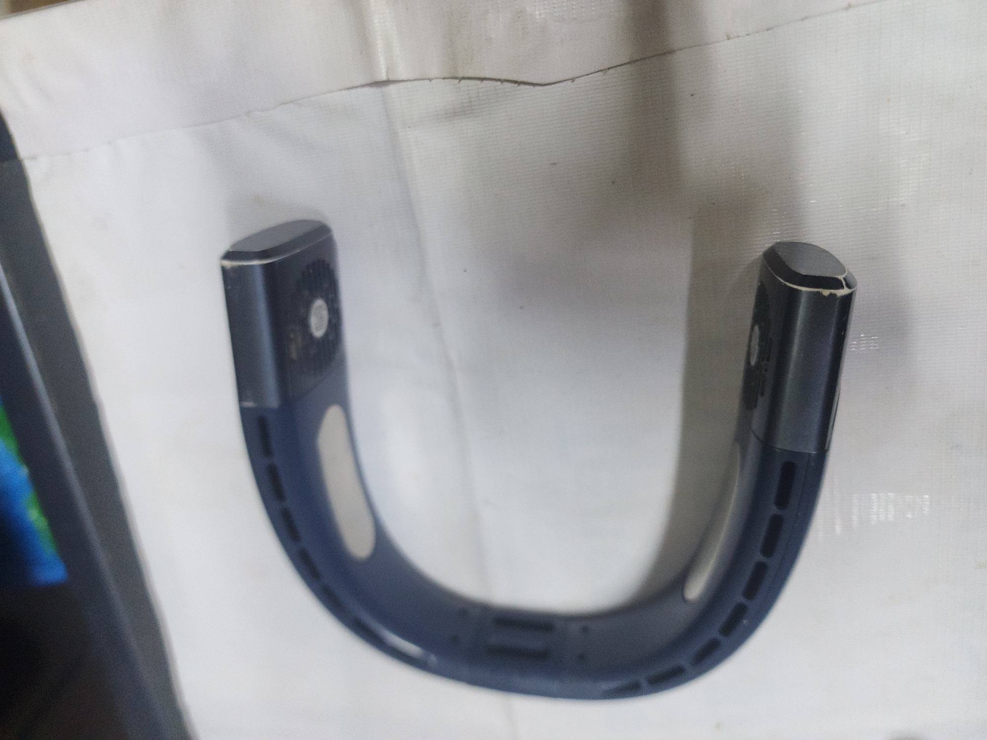 A dark blue, U-shaped personal fan rests on a white, textured surface. The fan has ventilation slots along its curved body and two black capped ends. One end of the fan has a circular vent, possibly for a motor or speaker. The fan is positioned on a white surface that appears to be fabric or paper. In the background, a dark, reflective surface is visible, with faint colors of blue and green. The lighting suggests an indoor environment. There are no people present in the image. The image quality is slightly blurry. It is not possible to determine the location in the city of Ushtobe, Kazakhstan, from the provided image.
